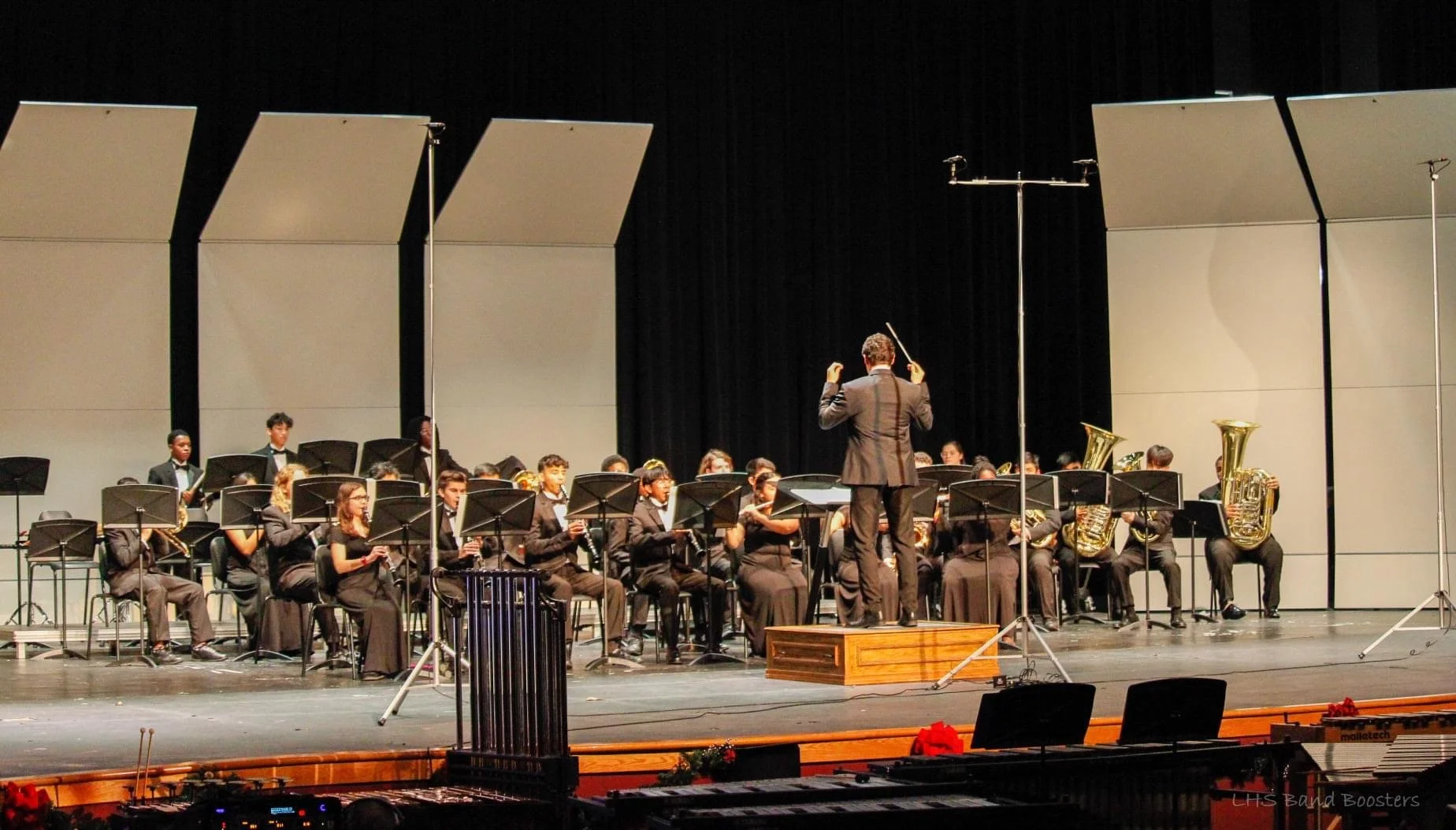 An orchestra performing on stage with a conductor standing on a wooden podium, facing the musicians. The orchestra includes string and brass instruments, arranged in rows. Large, vertical white and black stage panels background and music stands in front of musicians. Stage lighting focused on performers.