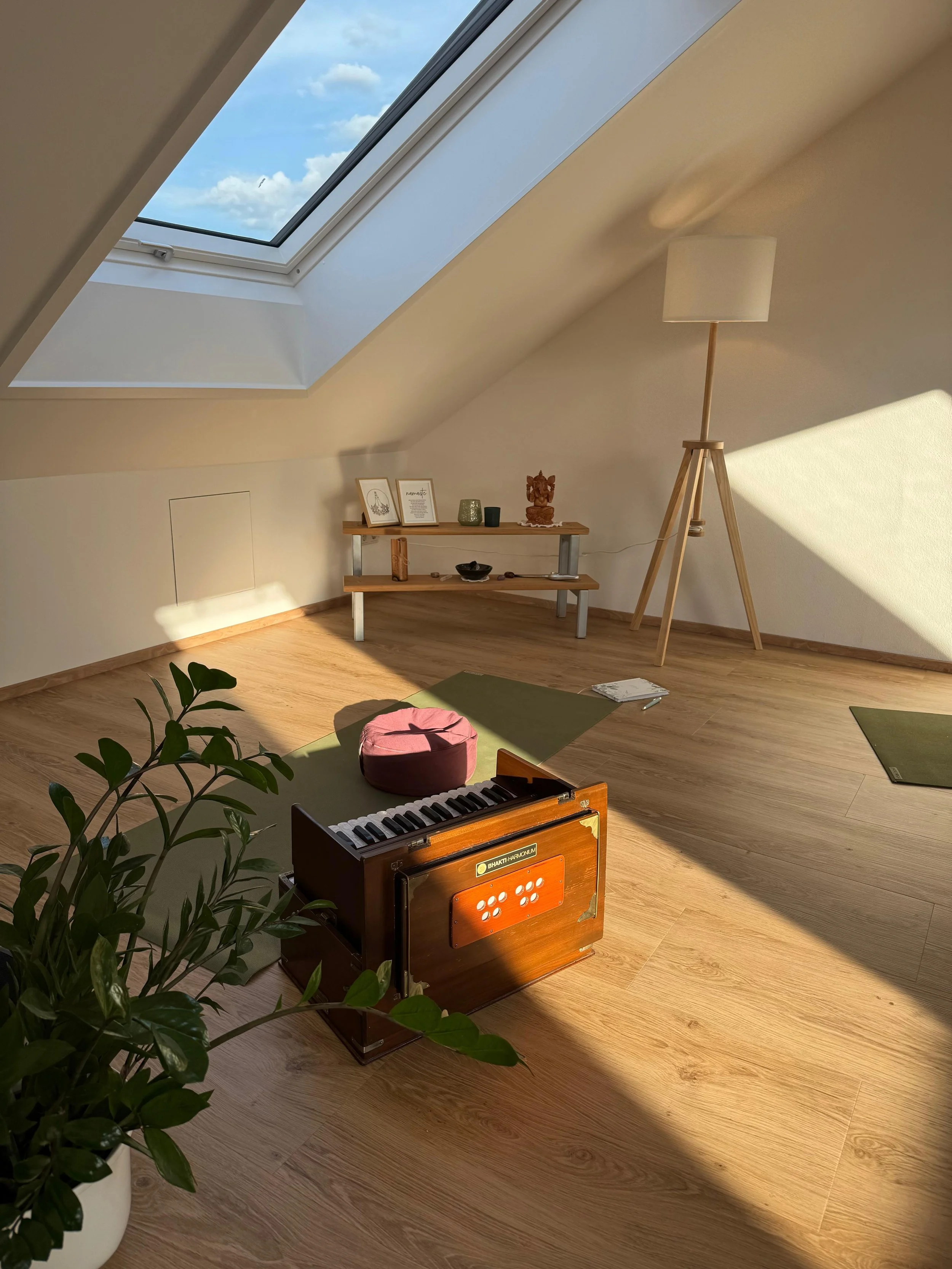 Ein heller Raum mit Holzfußboden, einer Stehlampe, einem Regal mit Dekorationen, einer Klavierklaviatur in einer Holzkiste und einem Pflanzenkübel mit grünem Blattwerk.