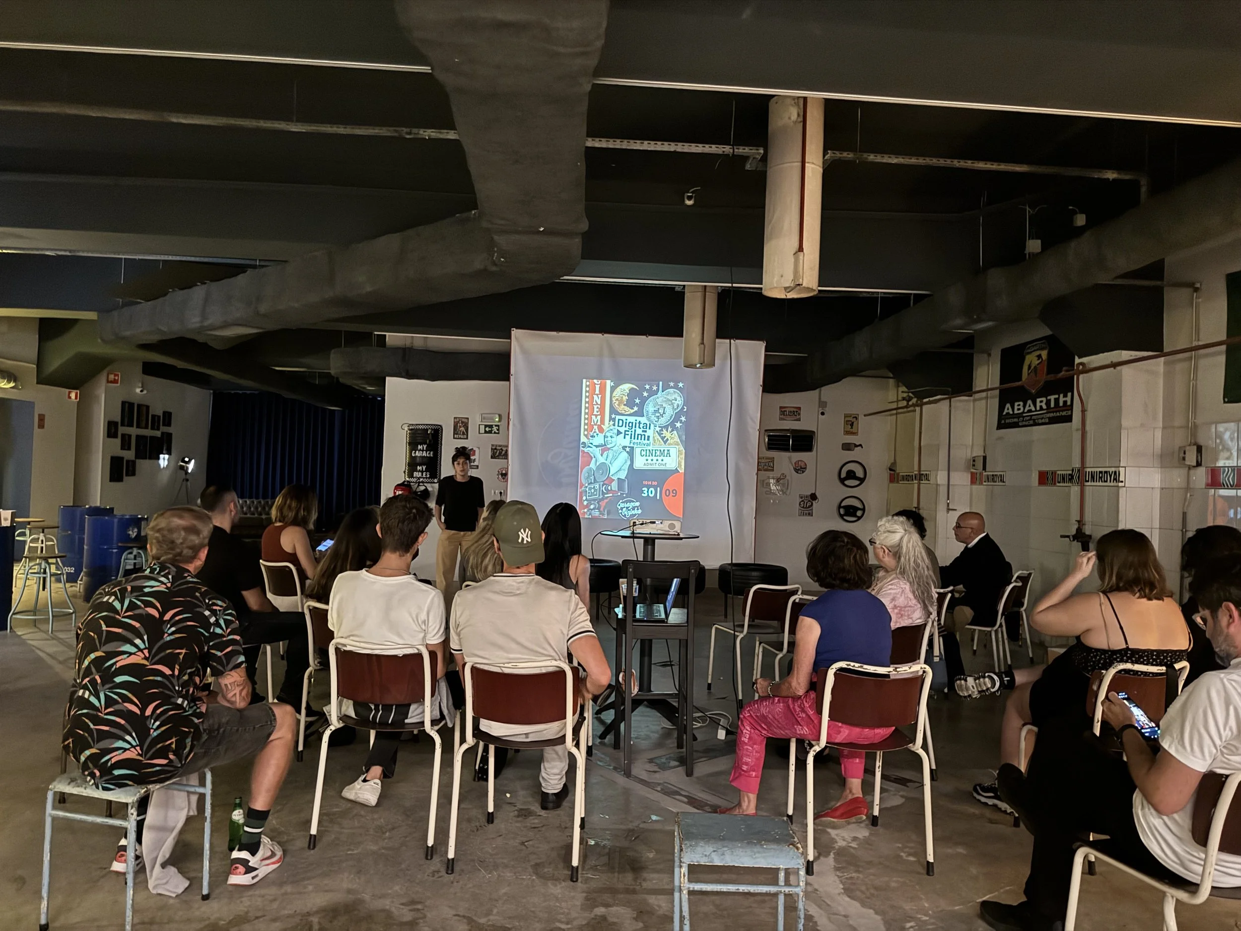 Pessoas assistindo a uma apresentação em um cinema digital, sentadas em cadeiras, diante de uma tela projetada com uma capa de filme e informações do evento.