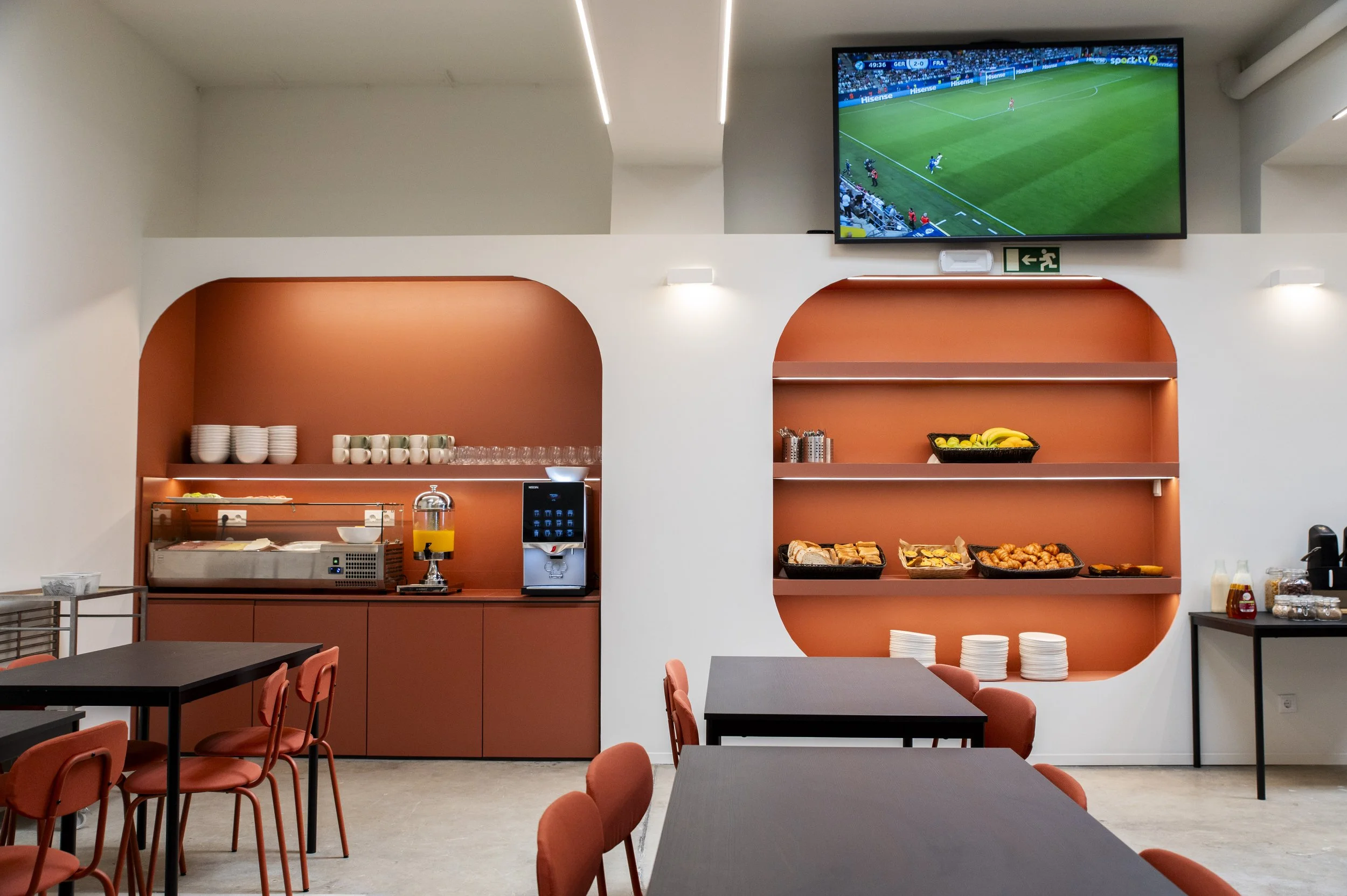 Sala de refeições com bancada de buffet, pratos de comida, uma televisão mostrando um jogo de futebol, e cadeiras laranja.