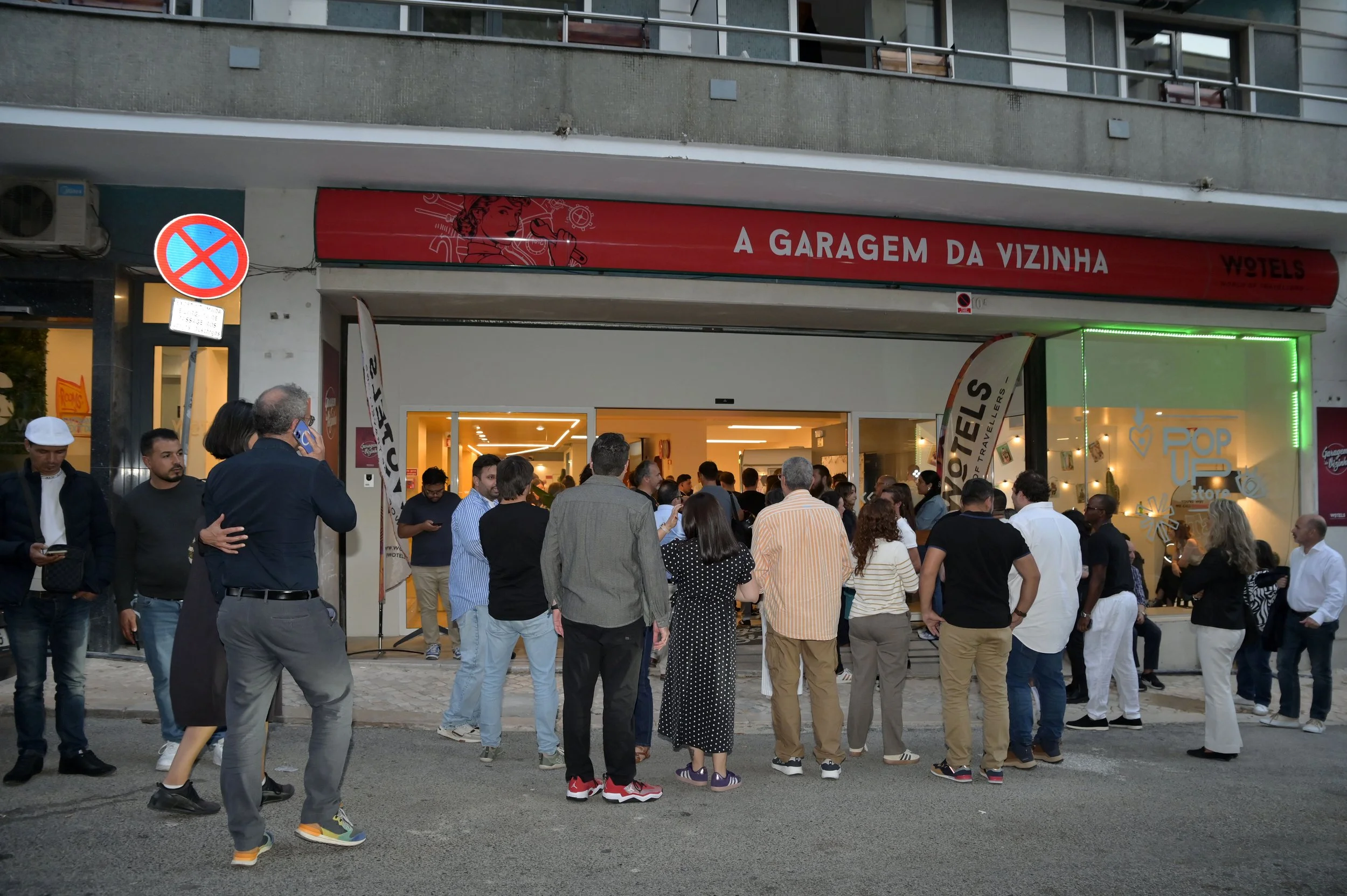 Fila de pessoas aguardando na entrada de um estabelecimento chamado 'A Garagem da Vizinha', um evento ou loja com iluminação interna amarelada e decoração moderna, localizado na rua em uma cidade brasileira.