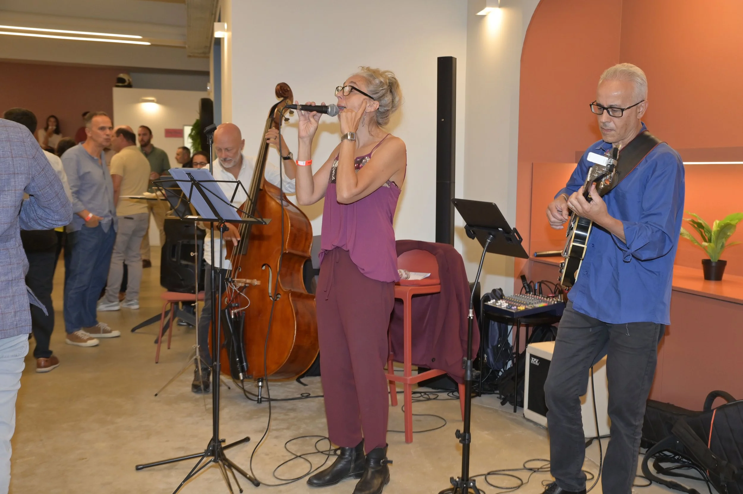 Casal de músicos tocando em evento social, incluindo uma mulher com violino e microfone e um homem com violão, com várias pessoas ao fundo.