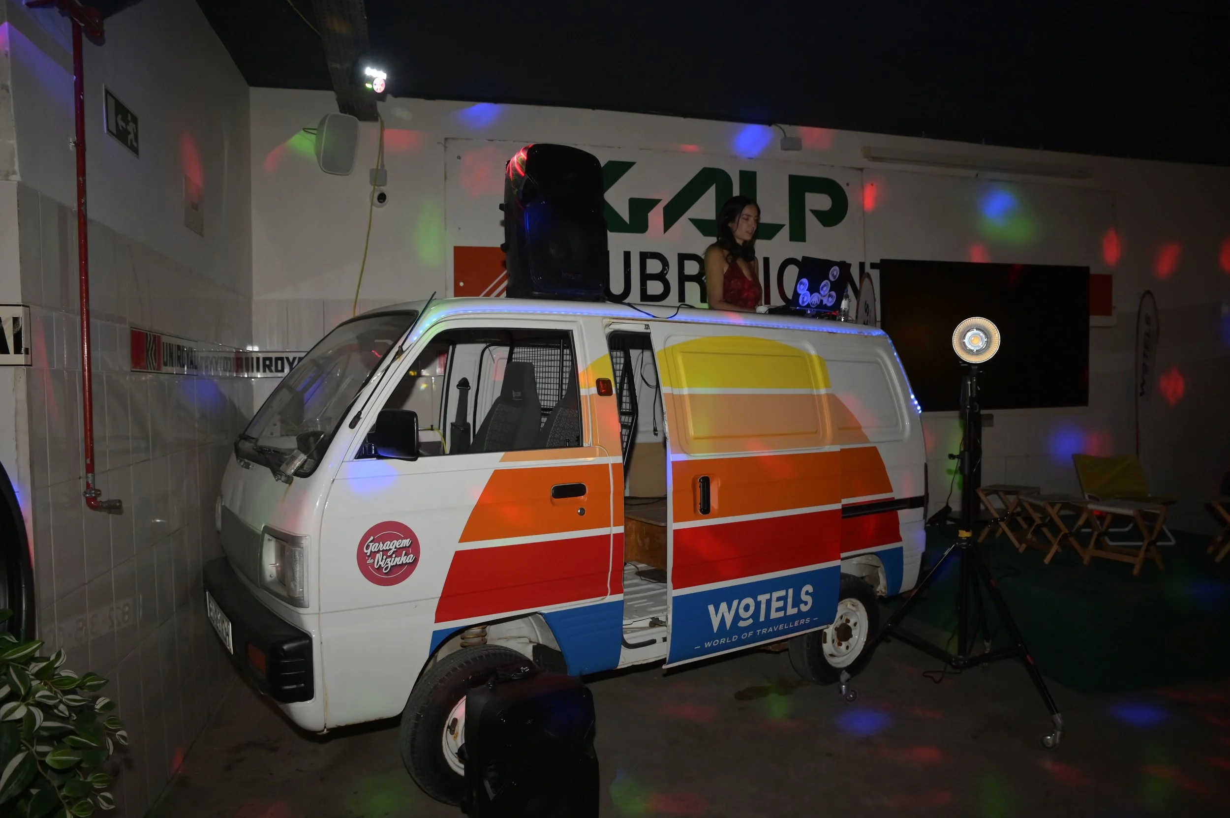 Mini van branca decorada com listras laranja, amarelas e azul, com anúncios de hotel e garagem na lataria, transformada em palco com DJ e som em um ambiente com luzes coloridas, cadeiras dobráveis e equipamento de iluminação.