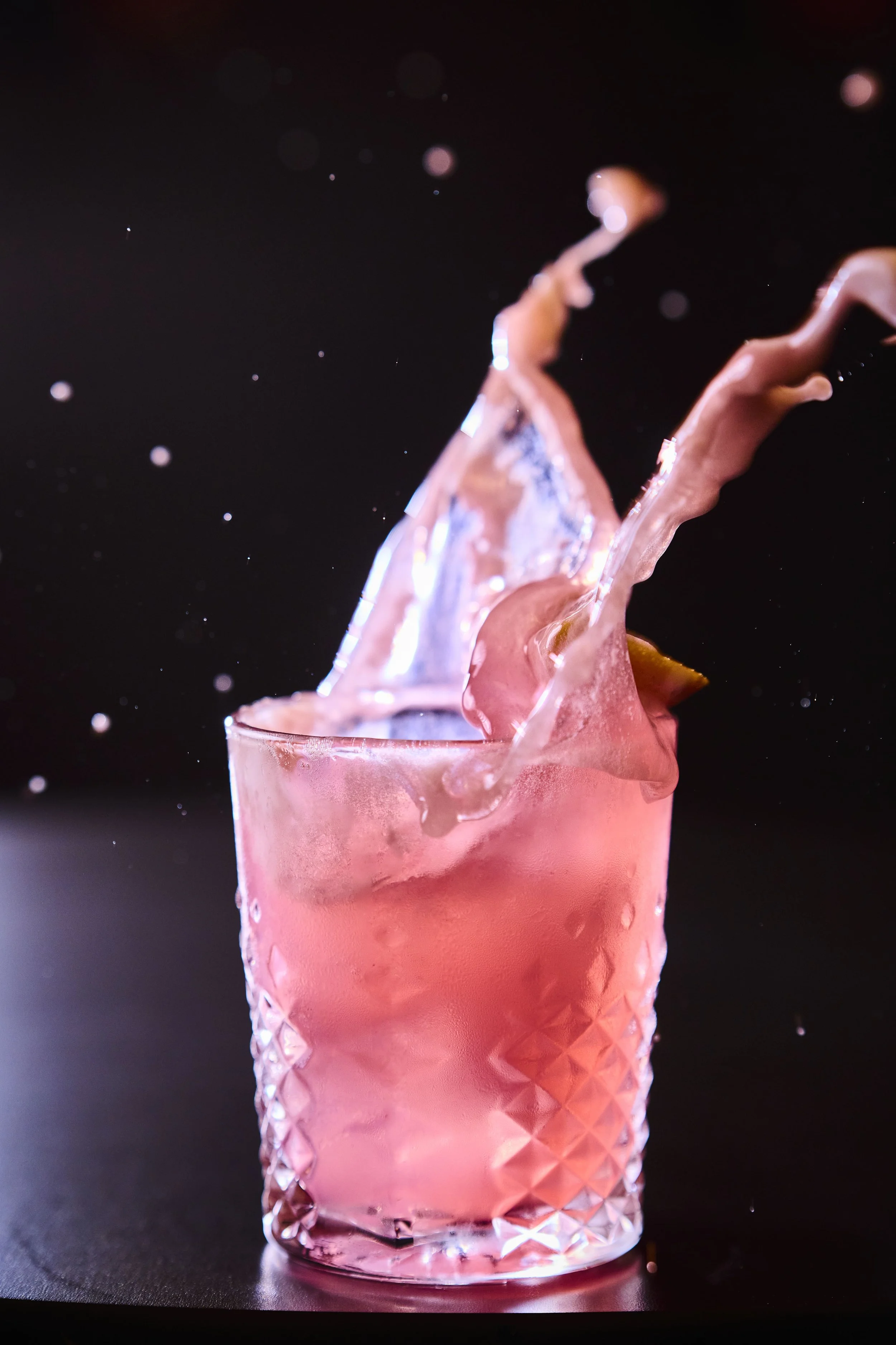 Copo de vidro com bebida rosa, com gelo, sendo derramada uma rosa de água. Fundo preto.