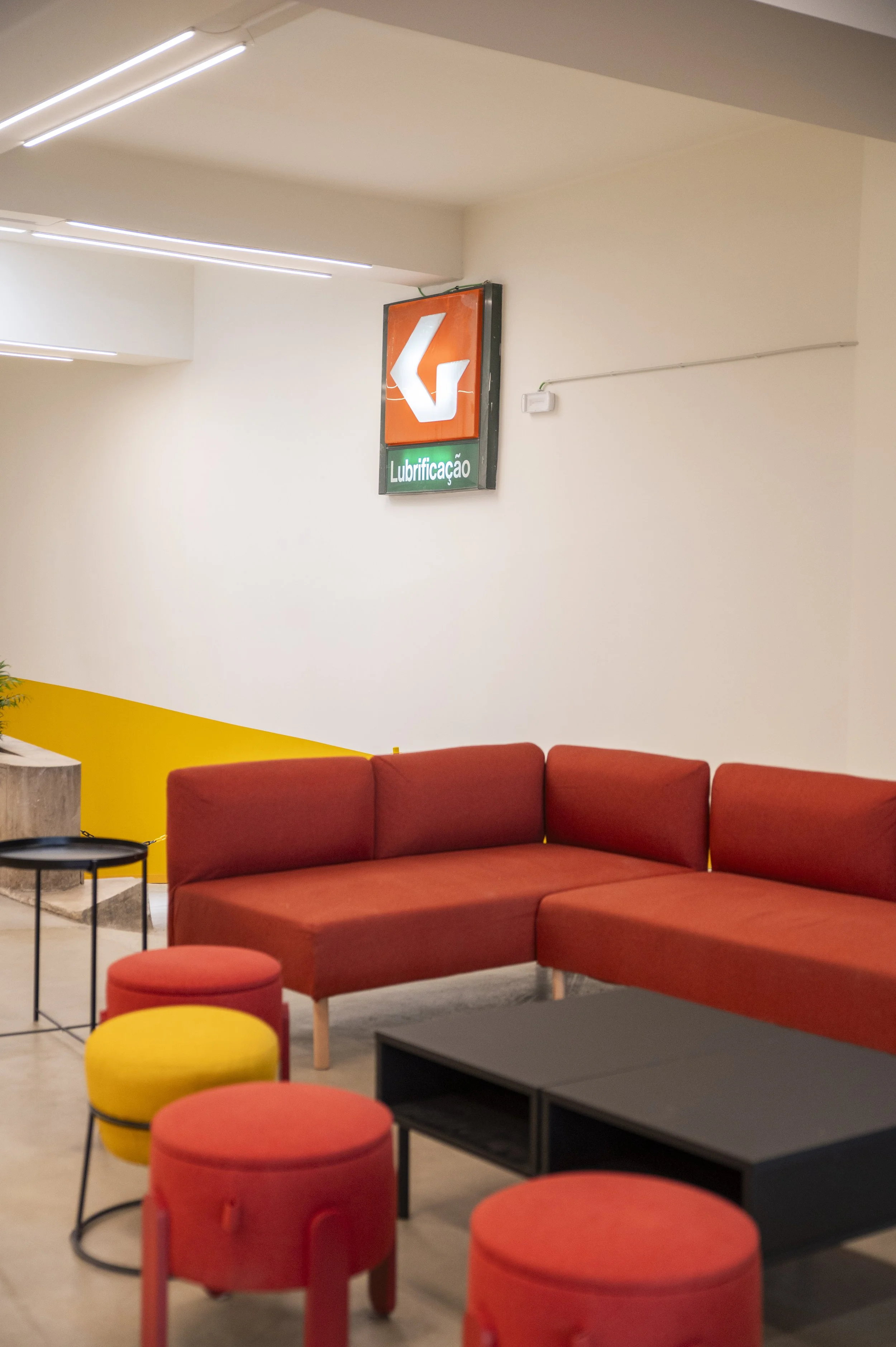 Sala de espera com poltrona vermelha, pufes amarelos e vermelhos, mesa baixa preta e iluminação moderna no teto. Na parede, sinalização de indicação de localização de lubrificação.