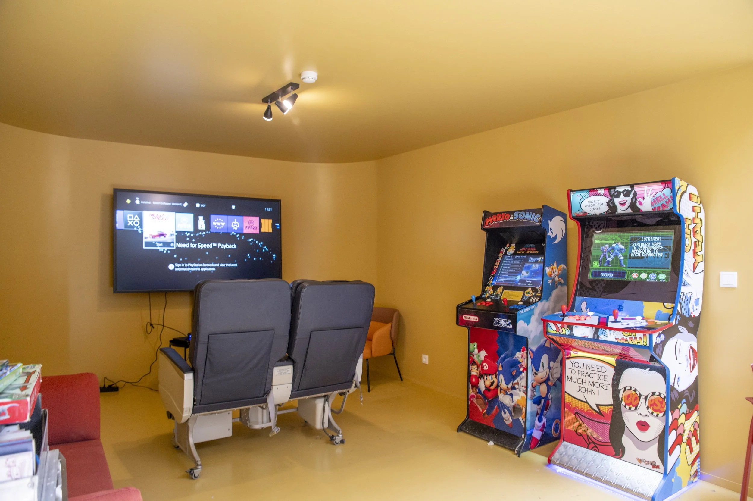 Sala de jogos com dois arcade games de Sonic e Mario, um sofá vermelho, cadeira de rodas com tela de televisão na parede exibindo interface de videogame e uma pequena mesa com livros.