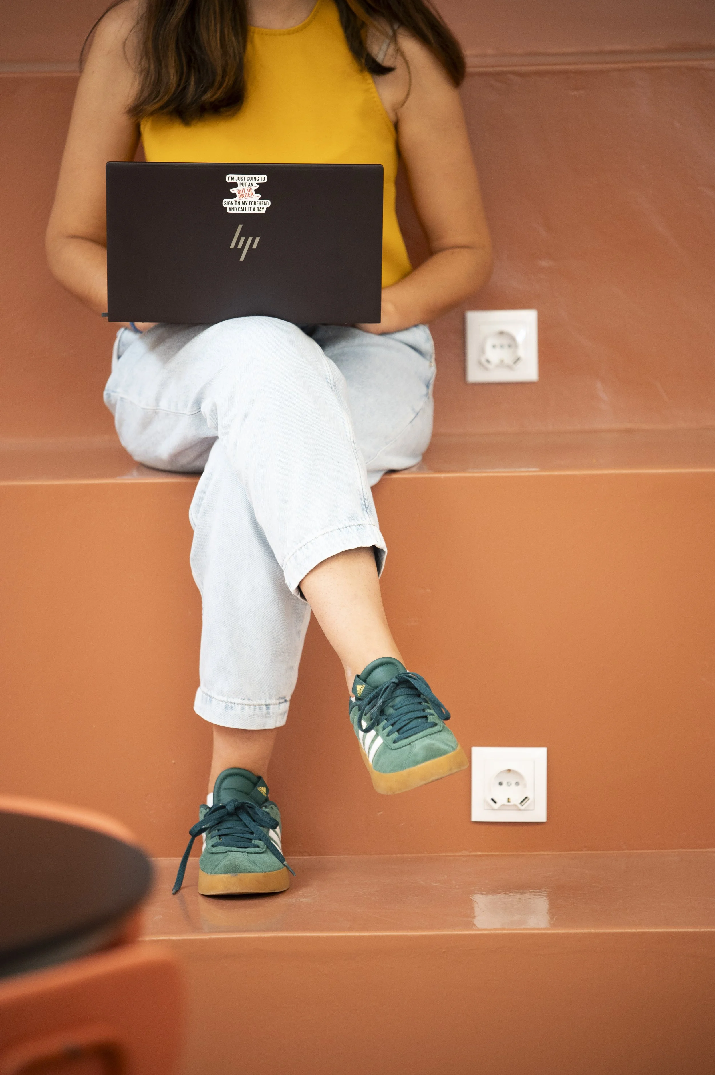 Pessoa sentada em escada com perna cruzada, usando tênis verdes, jeans e camiseta amarela, segurando um laptop preto com um adesivo na tampa.