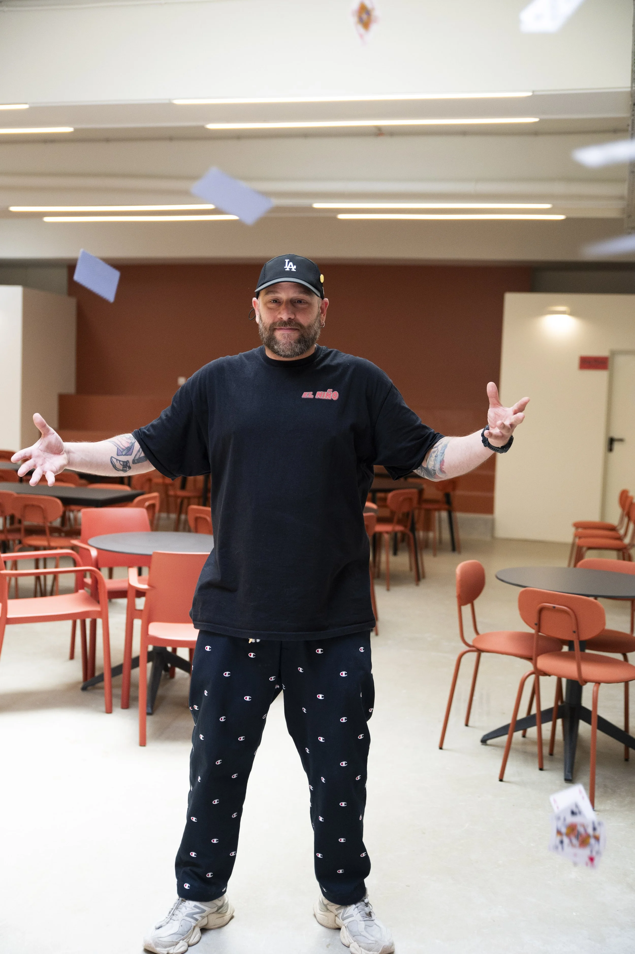 Homem com barba, usando boné preto, camiseta preta e calça de moletom com o logo da Champion, de braços abertos, em um espaço com mesas e cadeiras de cor vinho.