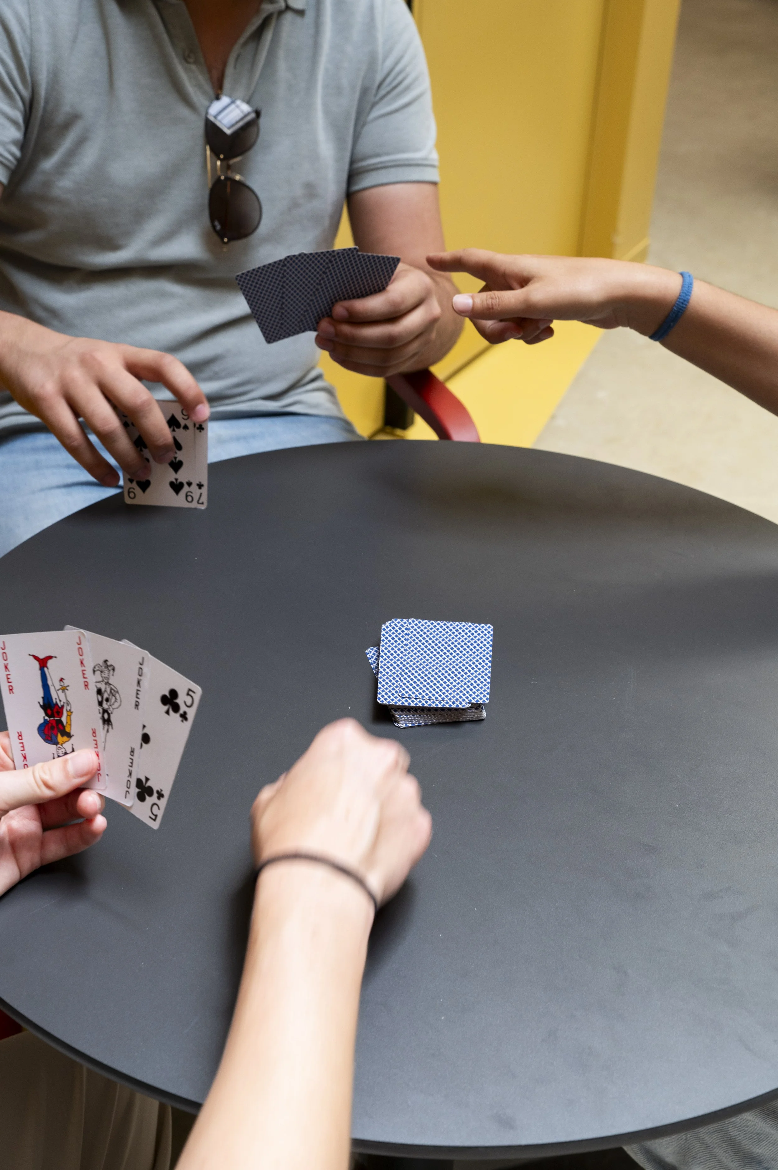 Três pessoas jogando cartas em uma mesa redonda preta, com uma pilha de cartas viradas para baixo no centro, em um ambiente com parede amarela ao fundo.