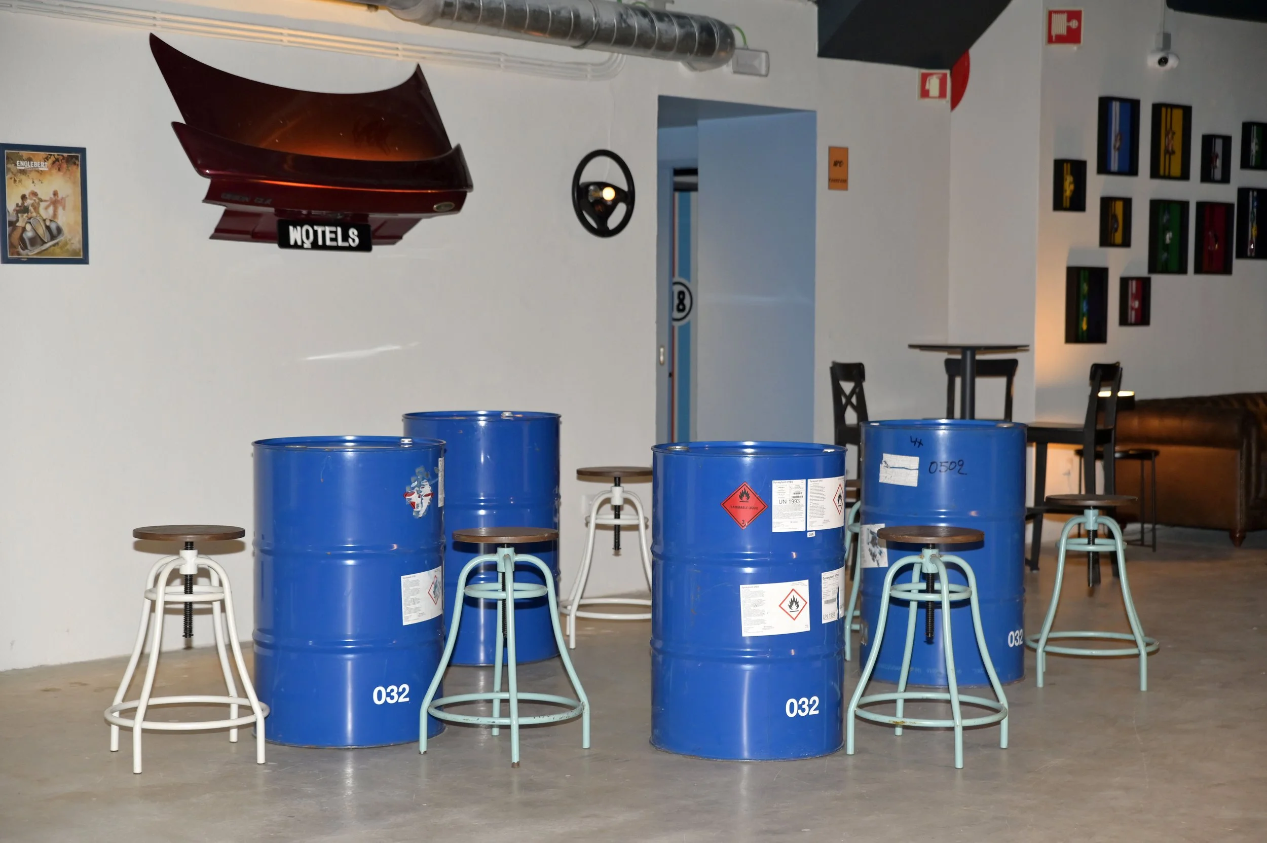 Vários barris de metal azul com símbolos de inflamável e peixes em uma área interna, com bancos de folhas de madeira empilhados em cima. Uma parede com quadros de cores diferentes ao fundo, e um painel de televisão antigo pendurado na parede.