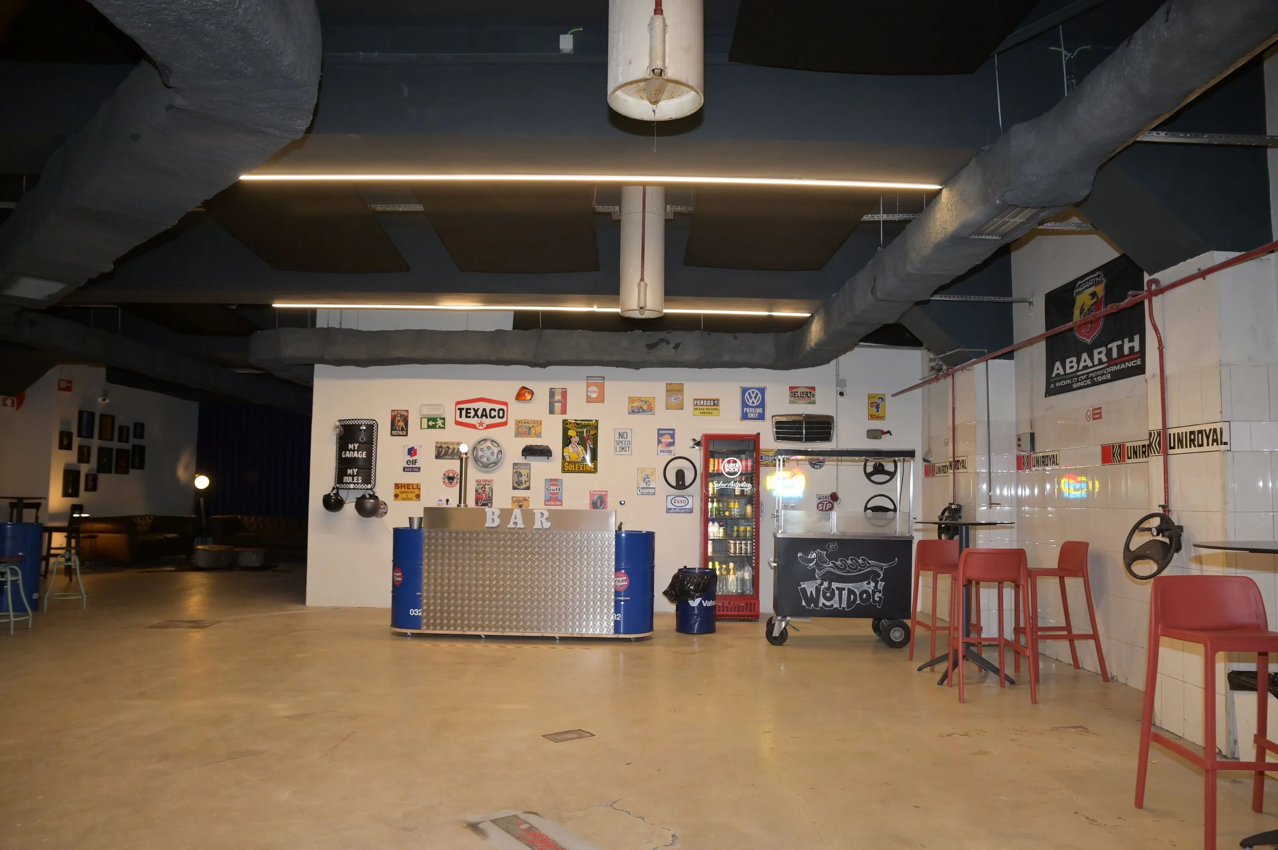 Bar com decoração temática de carros, com sinalizações de gasolina, docinhos e bebidas, cadeiras vermelhas e uma parede com várias placas e logos automotivos.