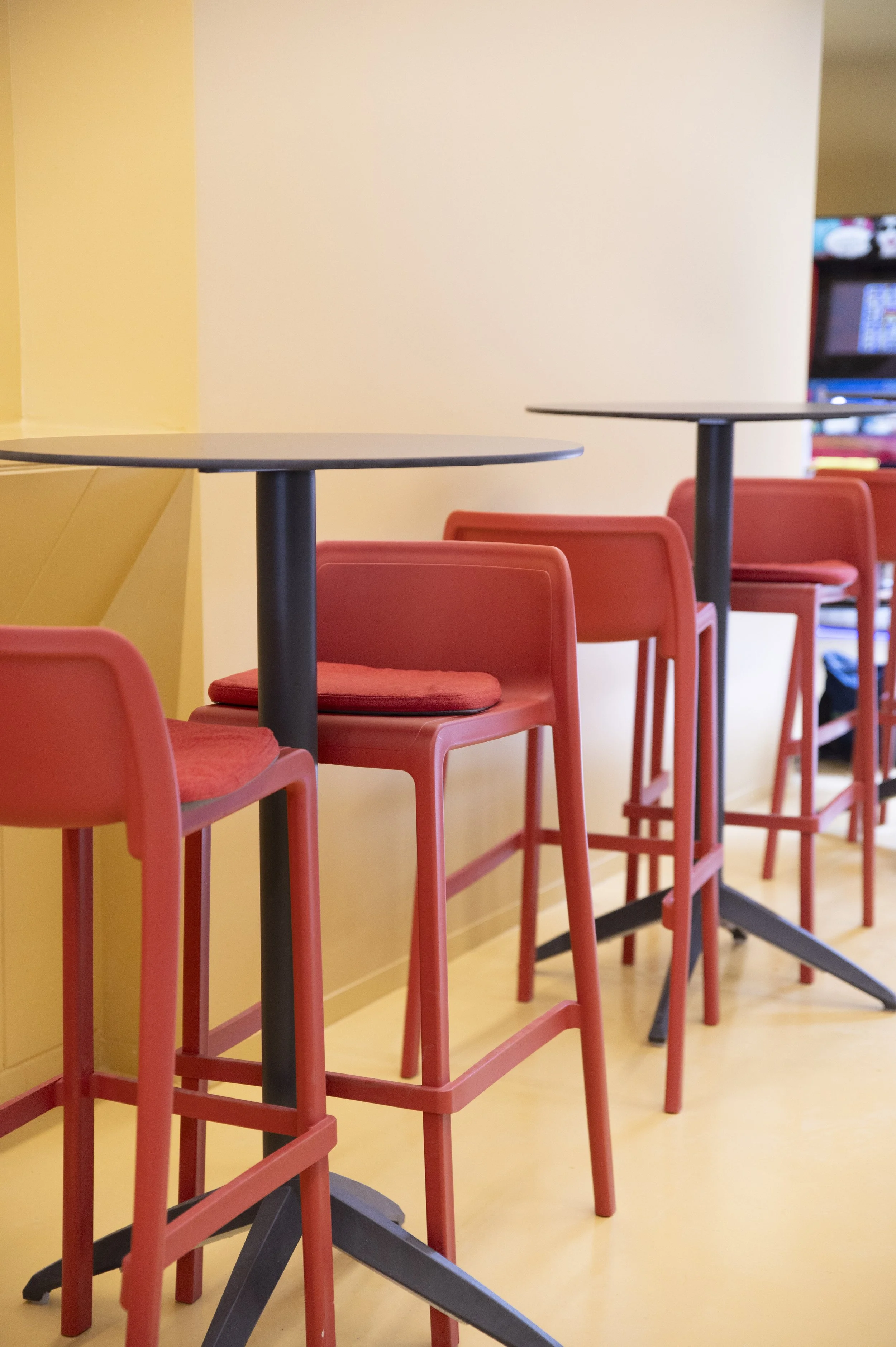 Cadeiras altas vermelhas empilhadas ao lado de mesas pretas em um ambiente interno com paredes amareladas.