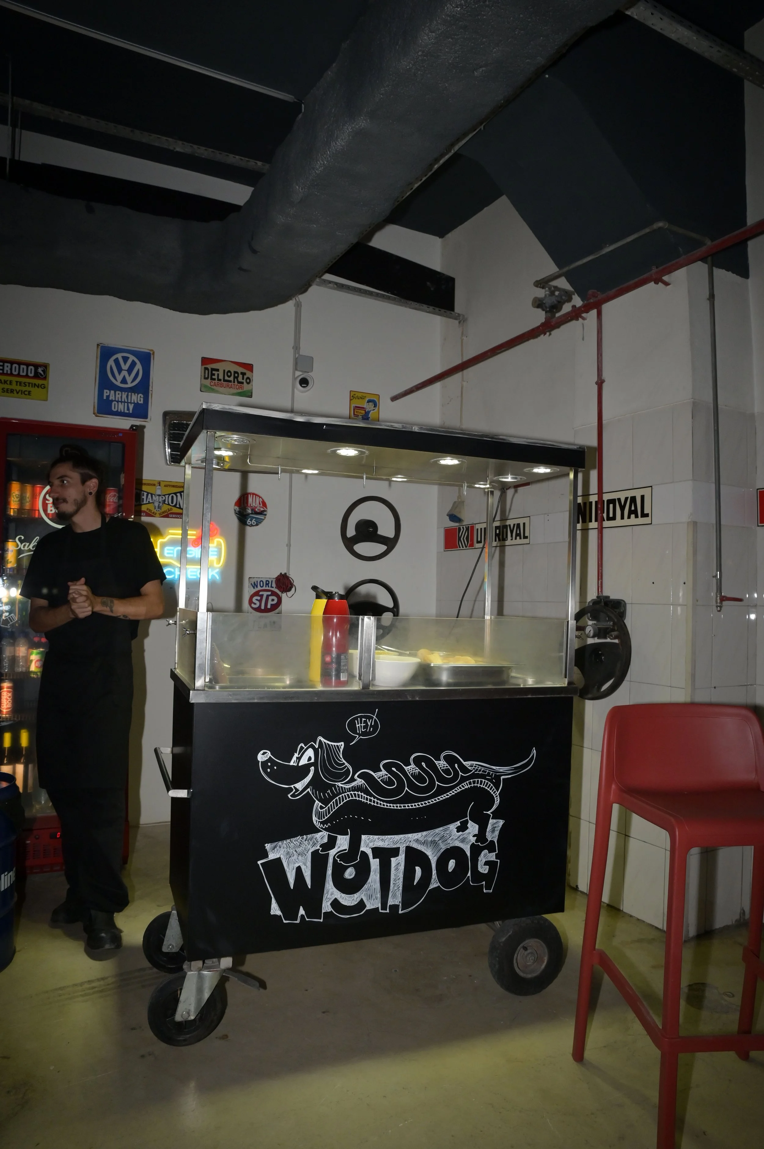 Carrinho de cachorro-quente com desenho de um cachorro com um chapéu de cowboy e a palavra 'WETDOG' na frente. Há ketchup e mostarda na bandeja, e uma pessoa ao lado.