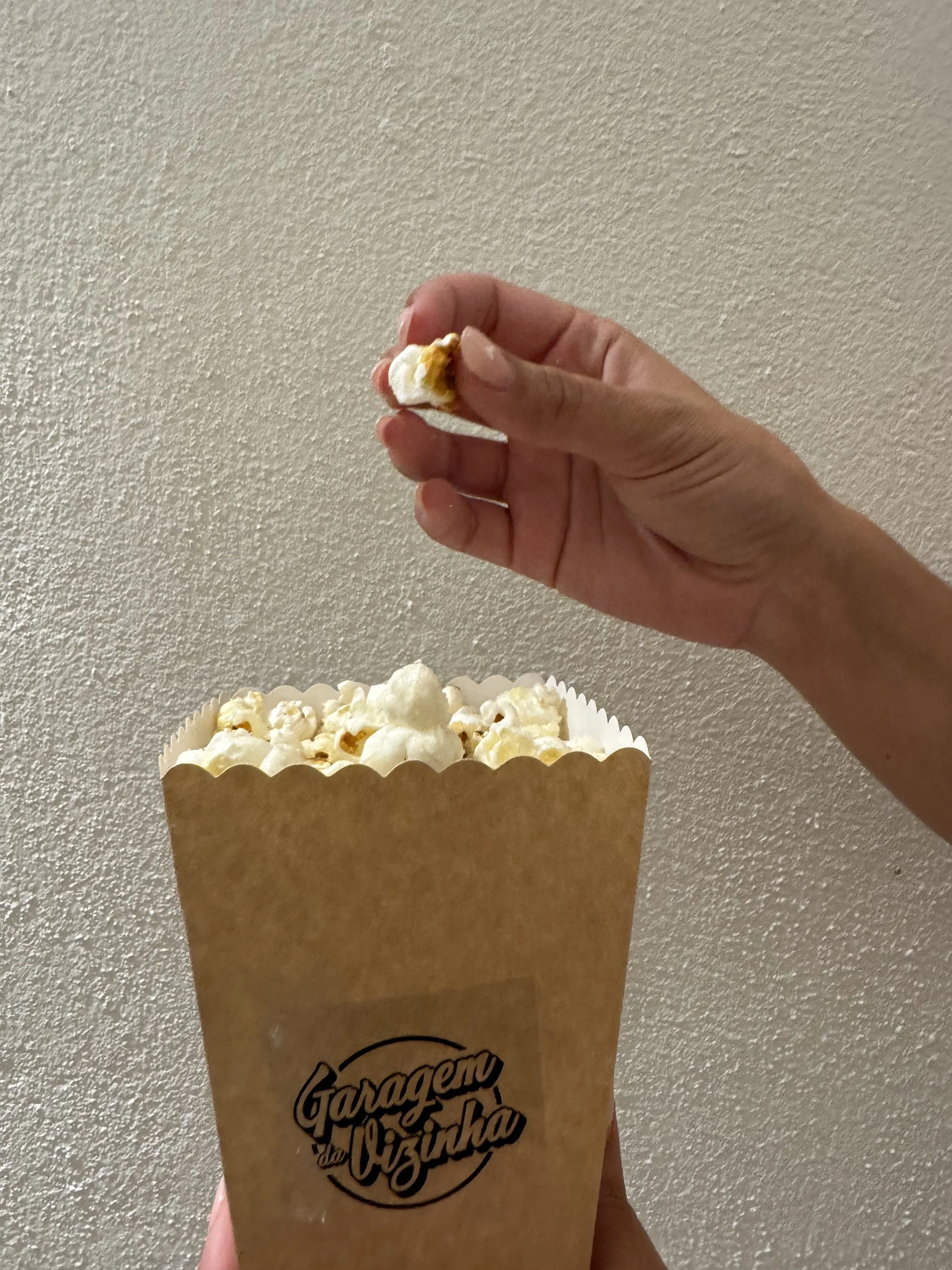 Pipoca na copo de papel com a marca 'Garagem da Vizinha', sendo segurada por uma pessoa contra uma parede