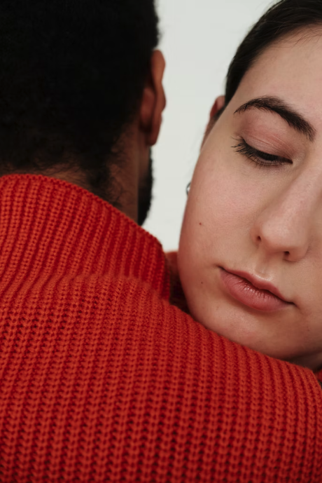 Close-up of a young woman with closed eyes, resting her head on a man's shoulder. The man is wearing a red sweater, and her facial expression is calm and serene.