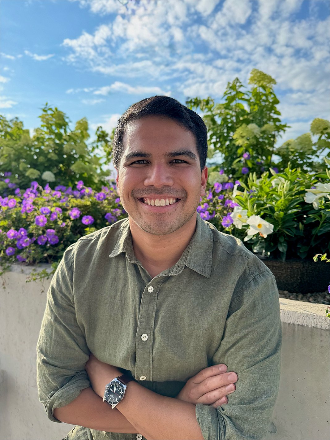A smiling male psychologist with dark hair, wearing a green button-up shirt and a watch, standing outdoors in front of a garden with purple and white flowers under a partly cloudy sky.