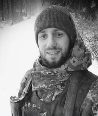 A man outdoors in a snowy forest, wearing a beanie, camouflage jacket, and scarf, smiling at the camera.