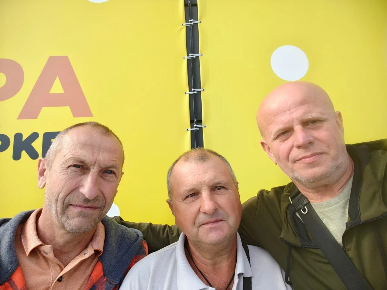 Three middle-aged men with light skin and short hair standing close together, taking a selfie in front of a yellow backdrop with some text and a white circle.