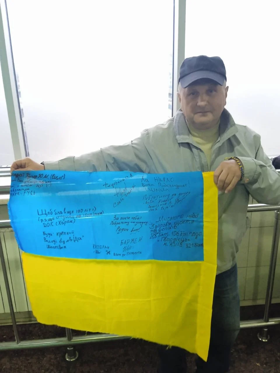 A person holding a Ukrainian flag with handwritten messages. The person is standing indoors, wearing a cap for protection from the cold or sun, a light-colored jacket, and a yellow shirt underneath. The flag is light blue and yellow, with various handwritten messages in blue ink.