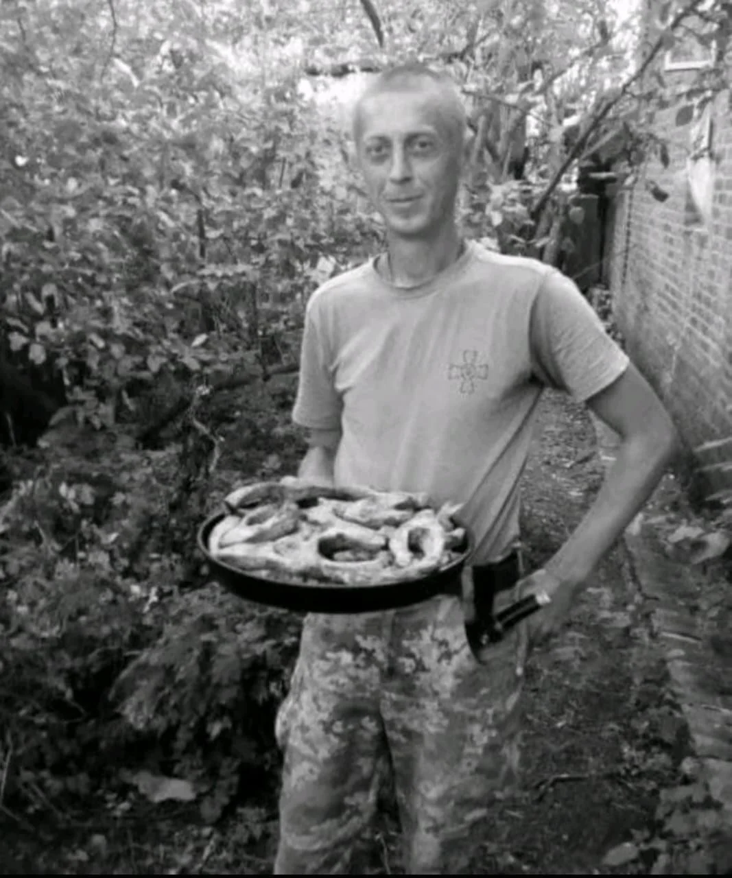 A man standing outdoors, holding a pan of grilled food, with trees and a brick wall in the background.