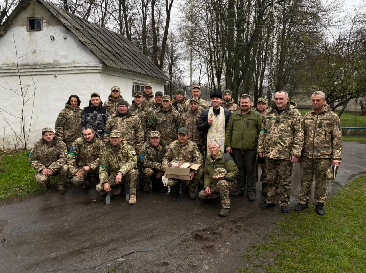 Group of soldiers in camouflage uniforms gathered outdoors near a white building with a gray roof. Some are standing, others kneeling, and two are holding packages.