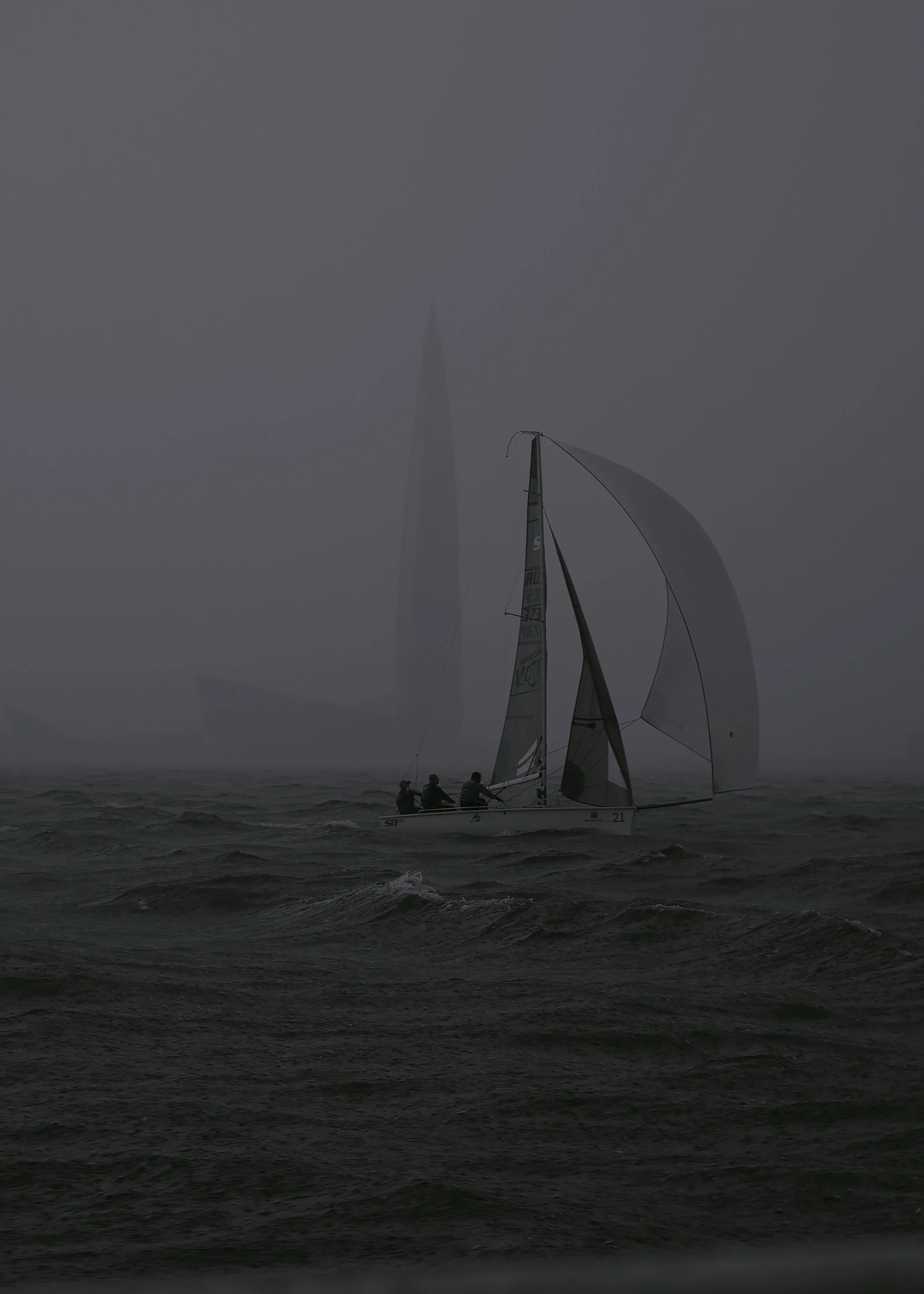 A sailboat with three people sailing through choppy waters of the Guft of Finland in foggy weather. In the background, the shape of the sailboat repeats the silhouette of the Lakhta center - the tallest building in St. Petersburg.