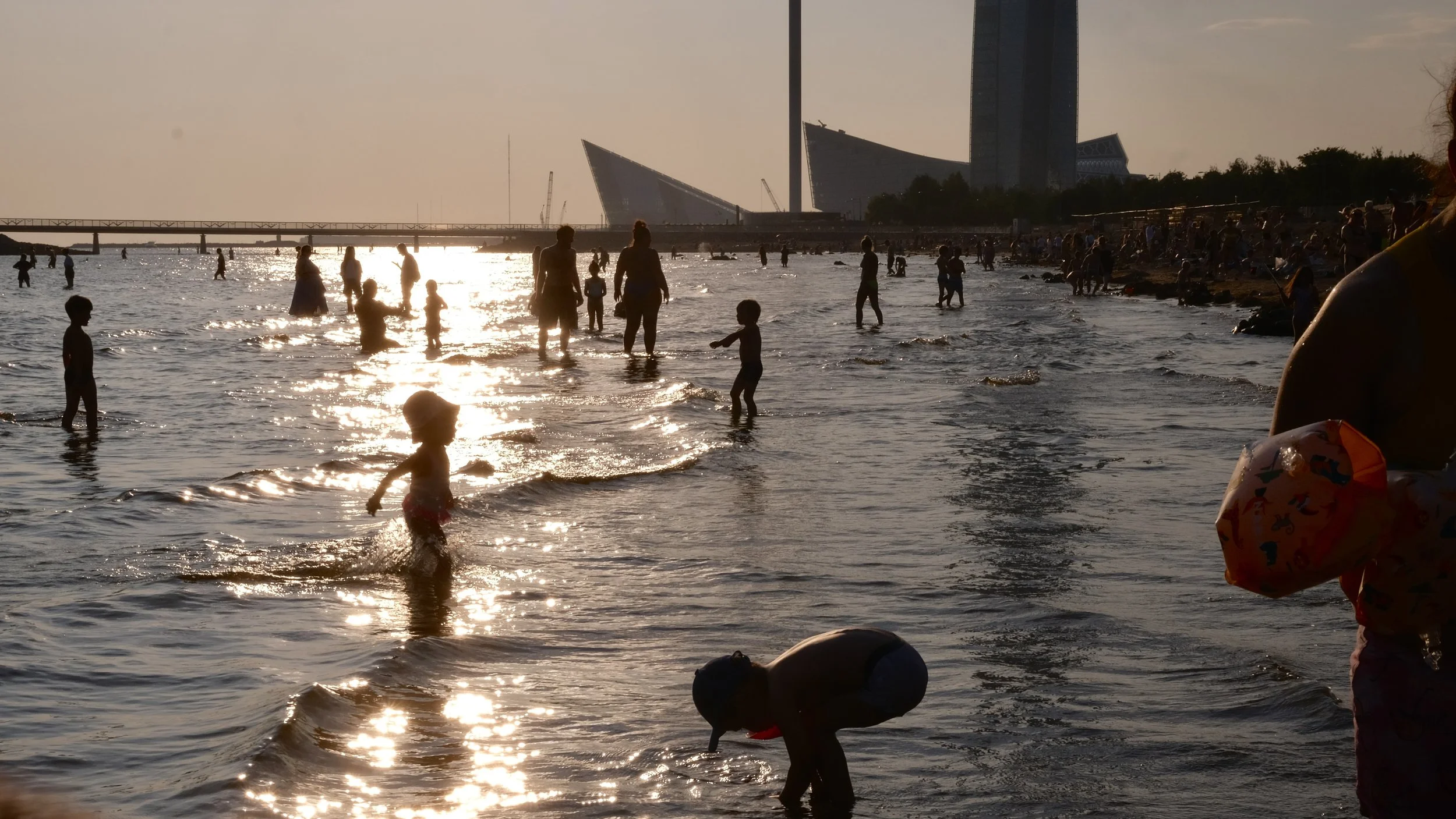 People enjoying a crowded beach during sunset, with children playing in the water and buildings in the background.