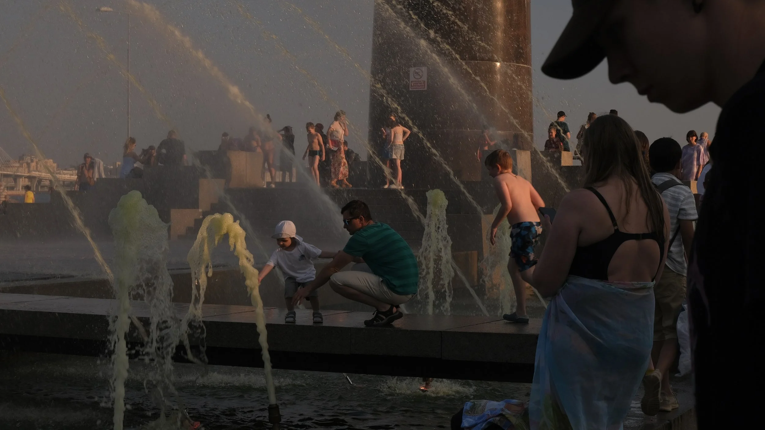 People enjoying a fountain with water jets during sunset. A woman holds a child, while others, including a man and a boy, interact with the water. More people are in the background around the fountain, with some on steps and others walking. The scene