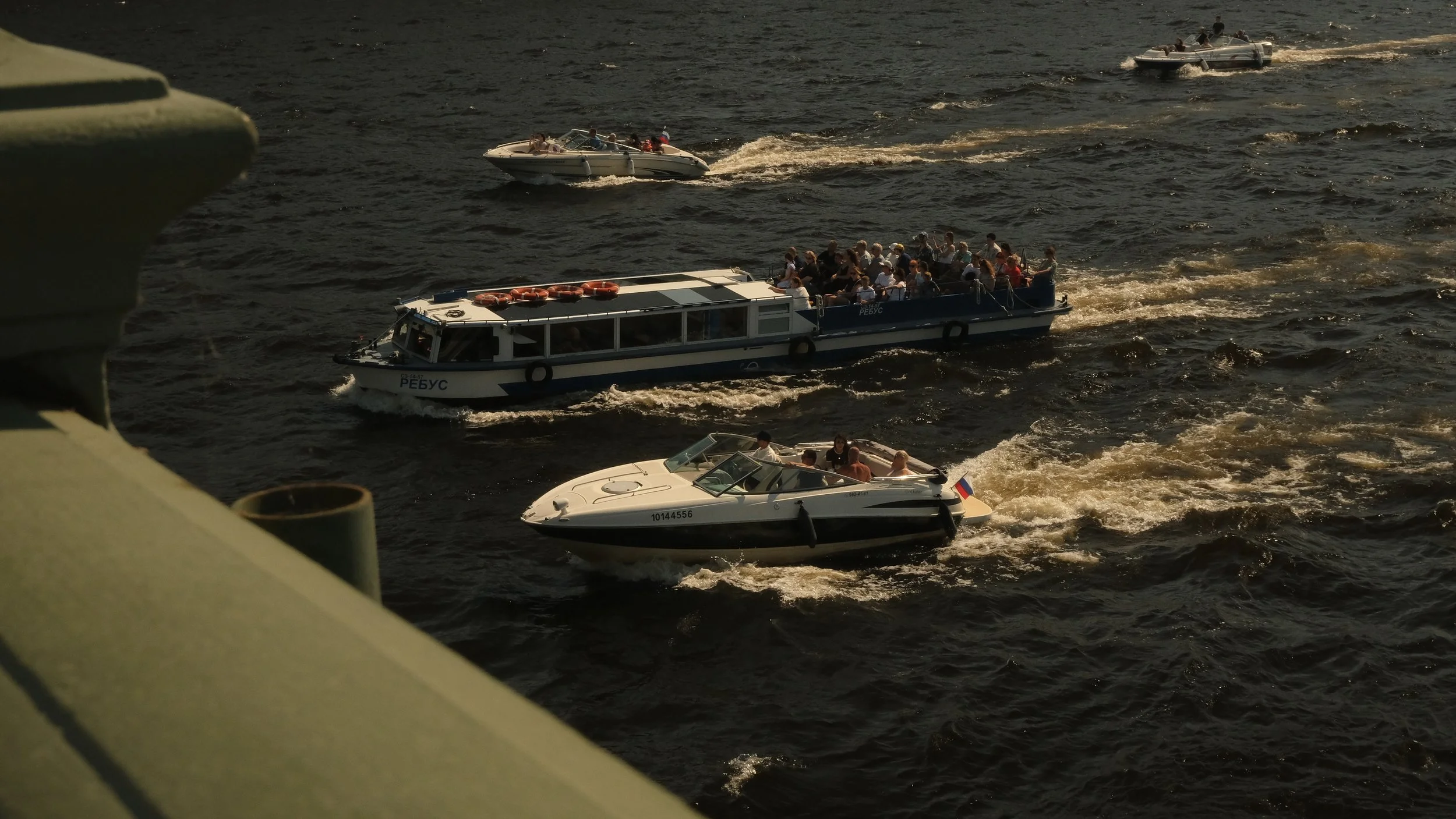 Multiple boats and ships on dark water, with people on board, seen from above from the bridge. Tourist season in St. Petersburg on the Neva River