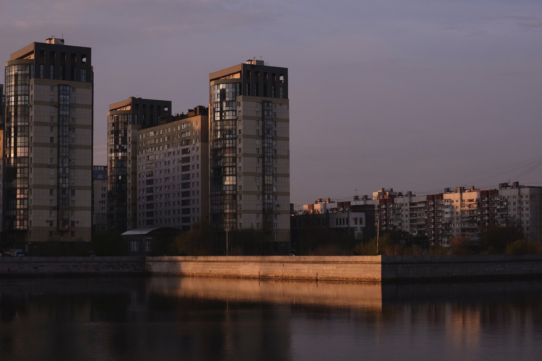 Cityscape with tall modern apartment buildings reflecting in a river during sunset.