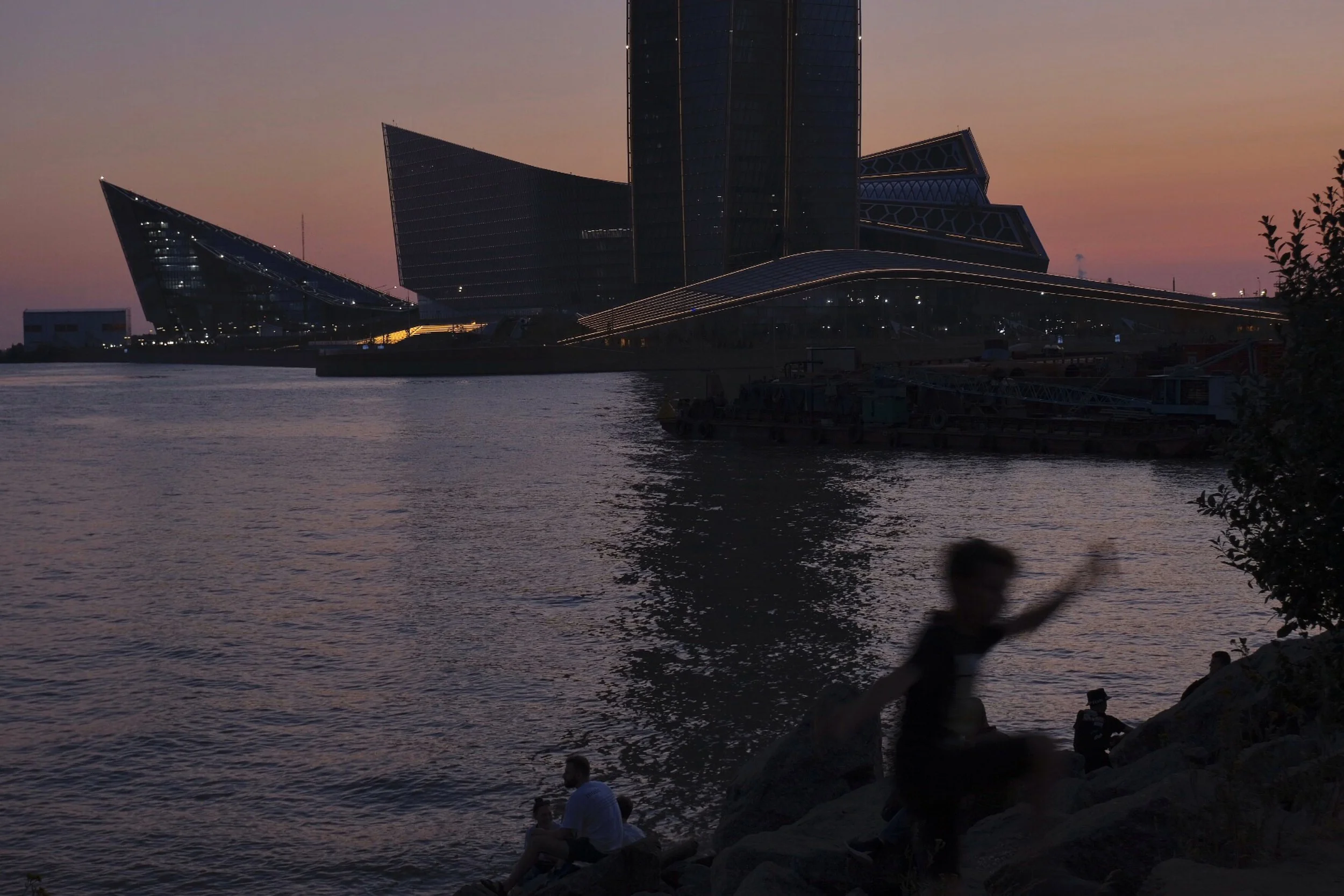 A waterfront cityscape at dusk with modern theater or office building with angular glass architecture, reflective water in the foreground, and a silhouetted person playing on rocks near the water. Other people sit along the shoreline, and the sky is 