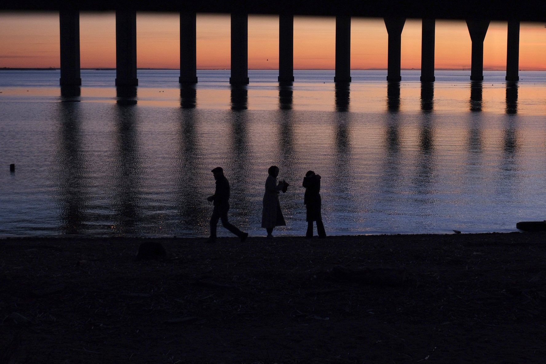 Silhouettes of three people walking along a beach at sunset, with a pier in the background and the water reflecting the evening sky.