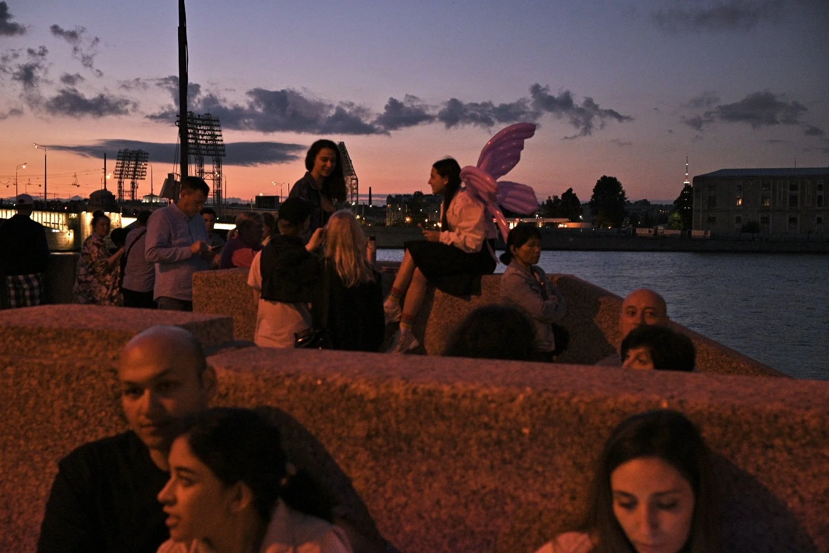 People gathered by a river at sunset, one person wearing pink fairy wings and a large pink bow, with a city skyline and cloudy sky in the background.
