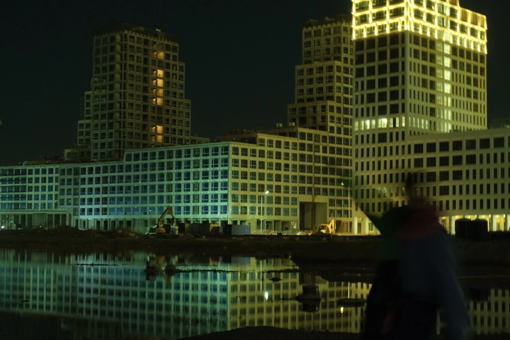 Nighttime construction site near water with illuminated multi-story buildings in the background and a blurred person in the foreground.