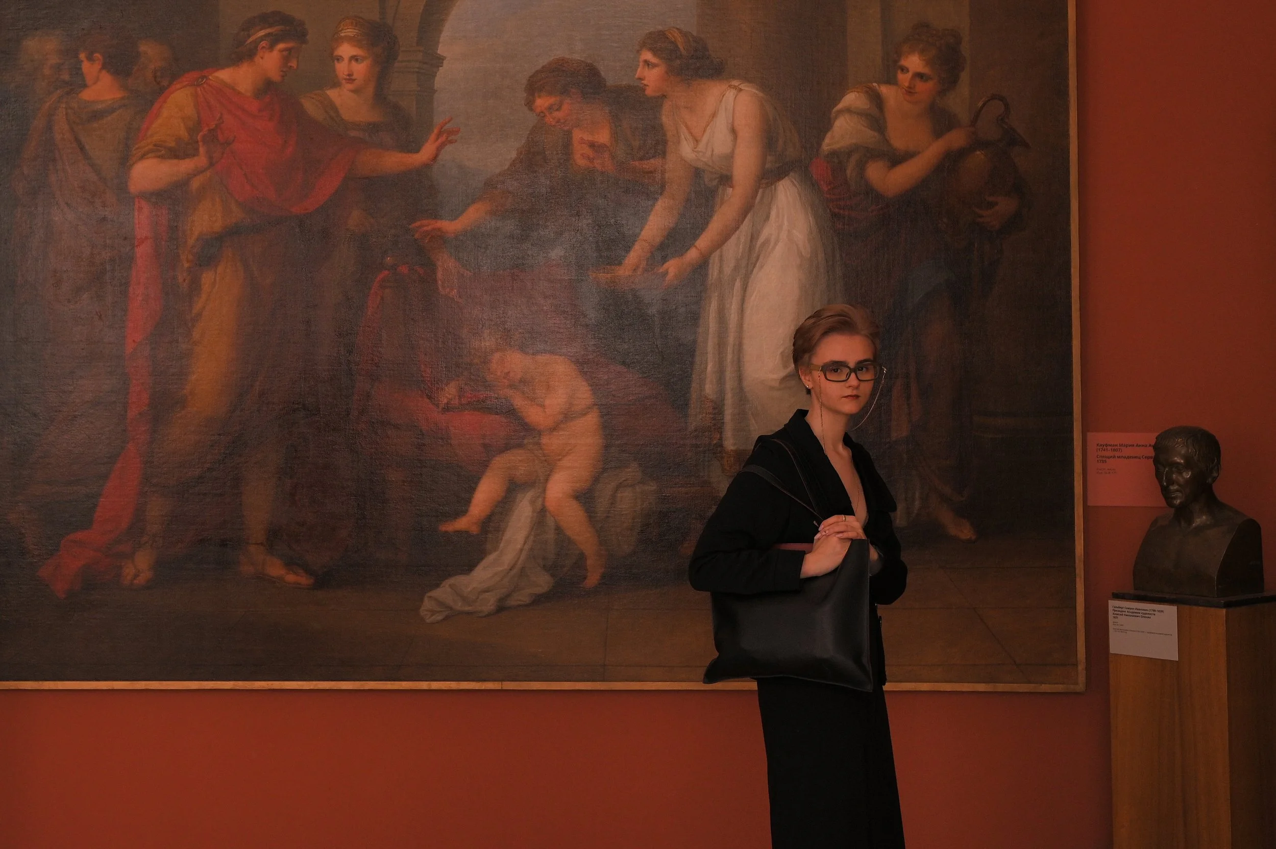 A woman wearing glasses and a black outfit standing in front of a large classical painting in an art gallery, with a bronze bust sculpture on a pedestal to the right.