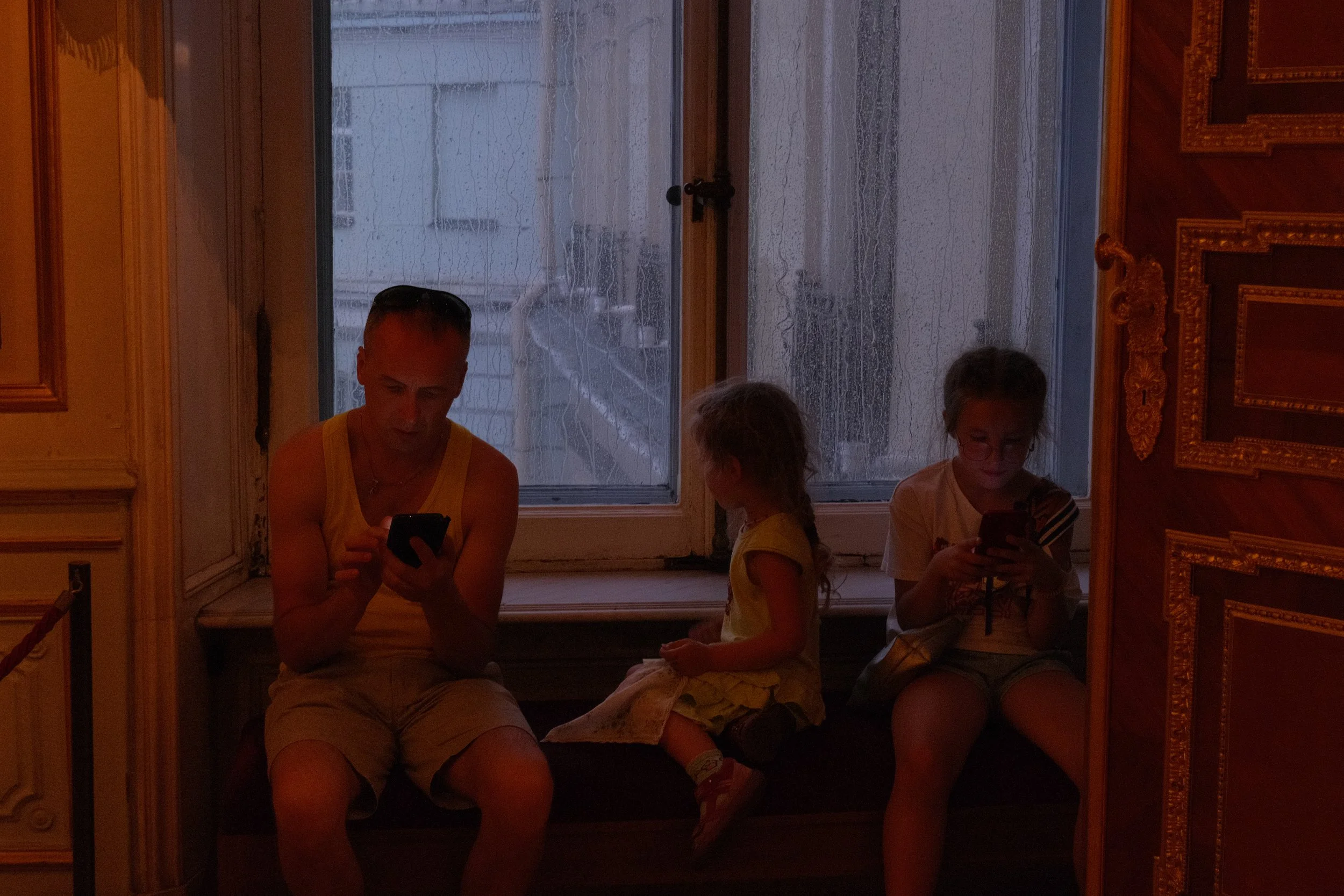 A man and two children sitting on a window seat, each looking at their smartphones inside a dimly lit room with ornate wooden paneling and a rain-covered window in the background. Street photography in Saint-Petersburg museums