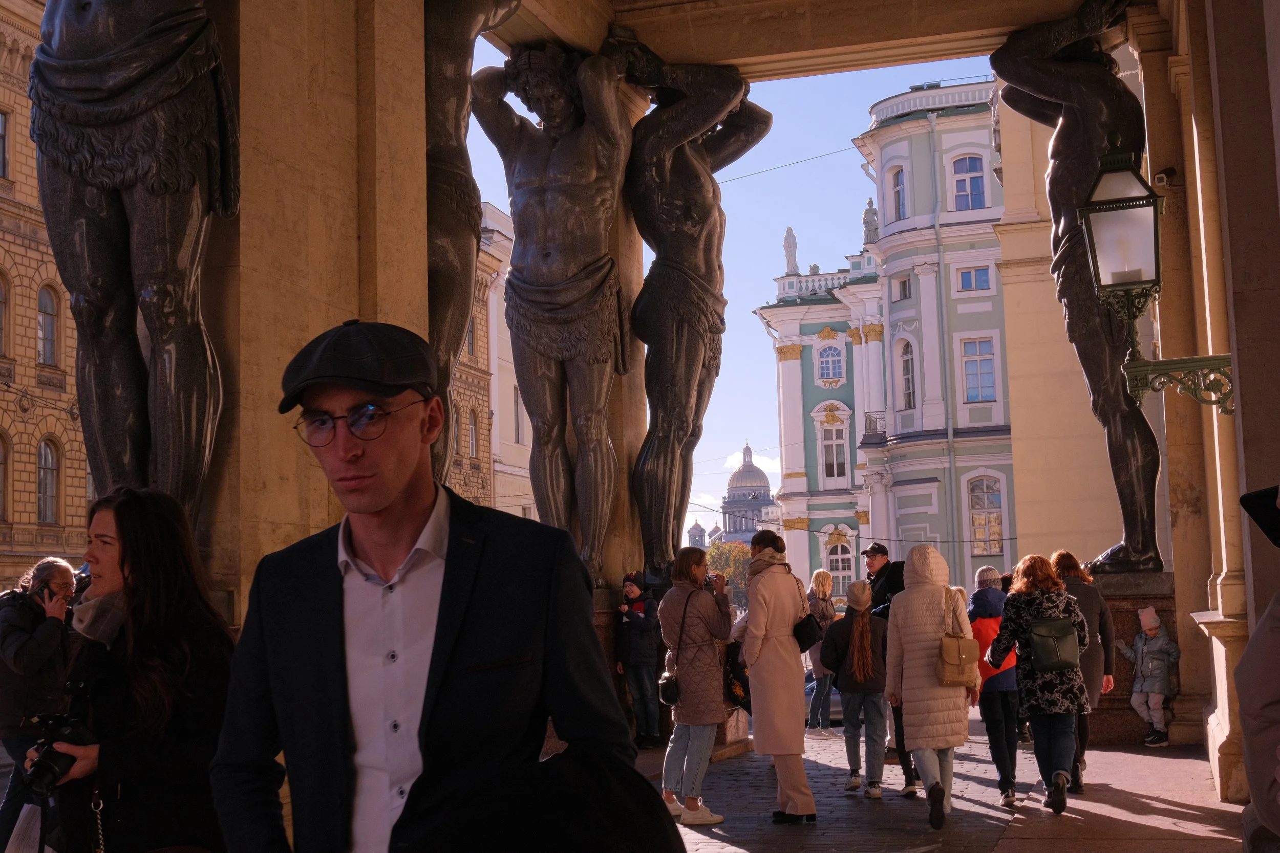 People walking and talking near the portico with Atlanteans at the entrance to the Hermitage. In the foreground is a man in a cap and glasses with a pensive look, and behind him, Atlanteans hold the portico.