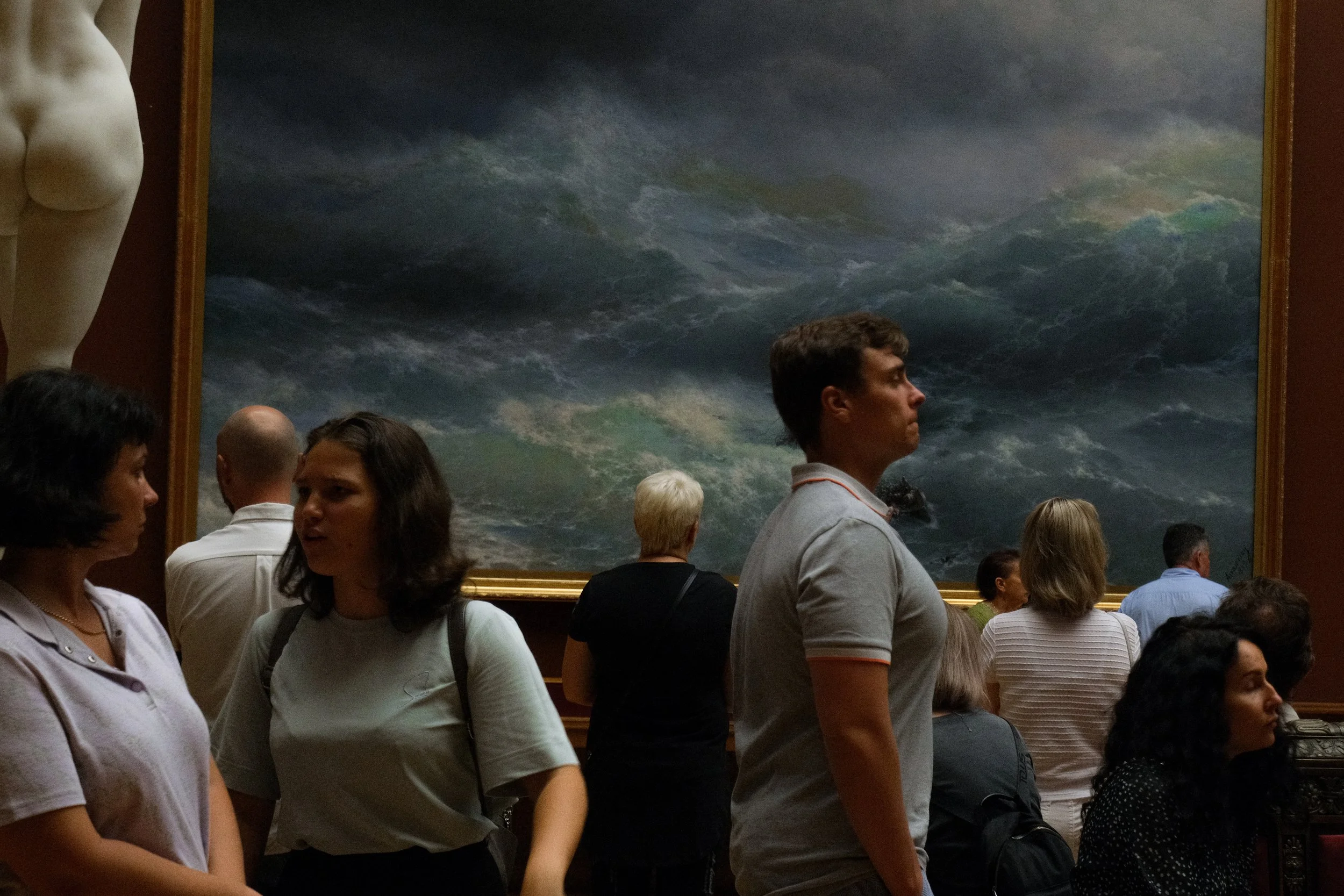 People observing an ocean scene painting at an art gallery, with a statue visible on the left.