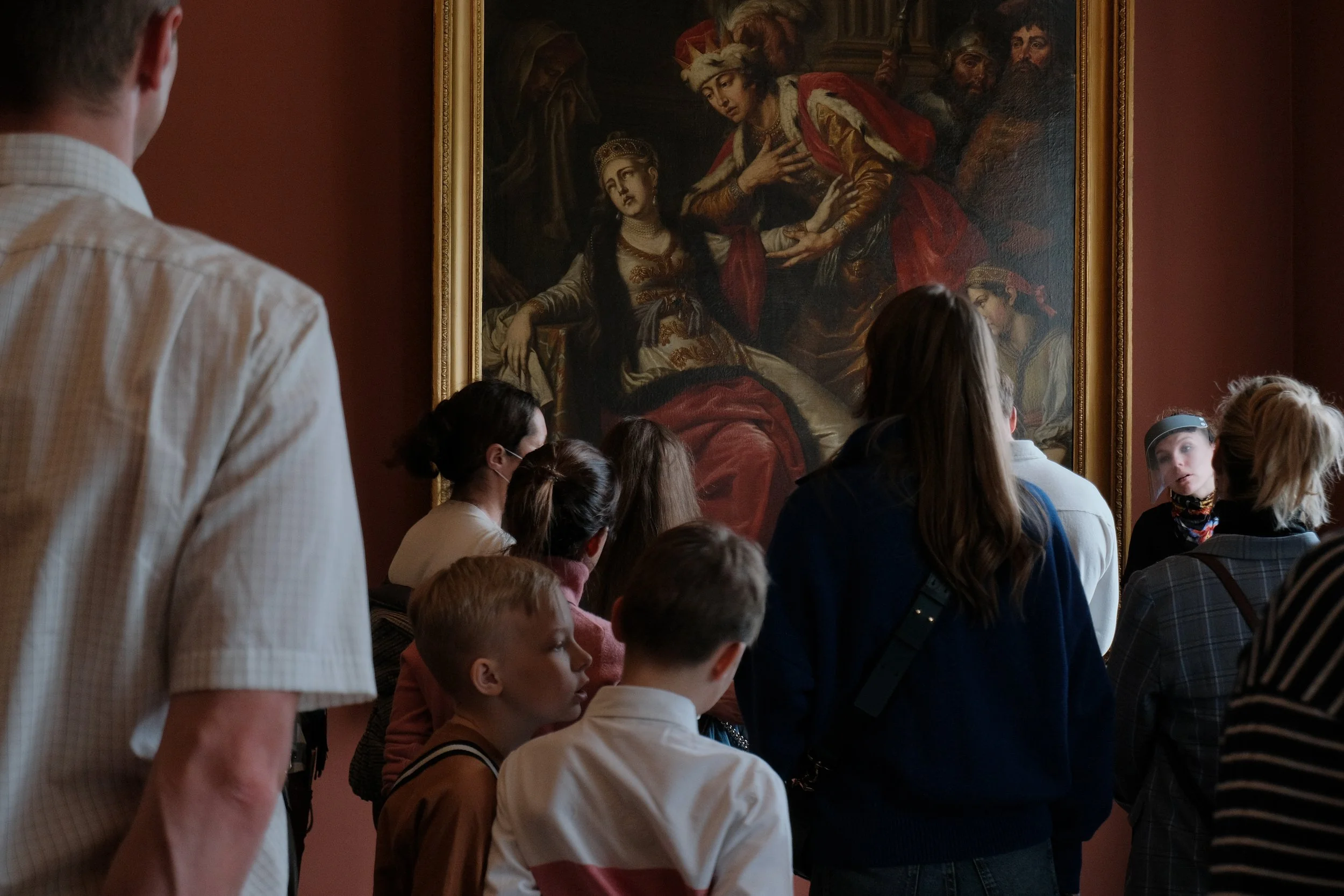 Group of children and adults viewing a large classical oil painting in an art gallery.