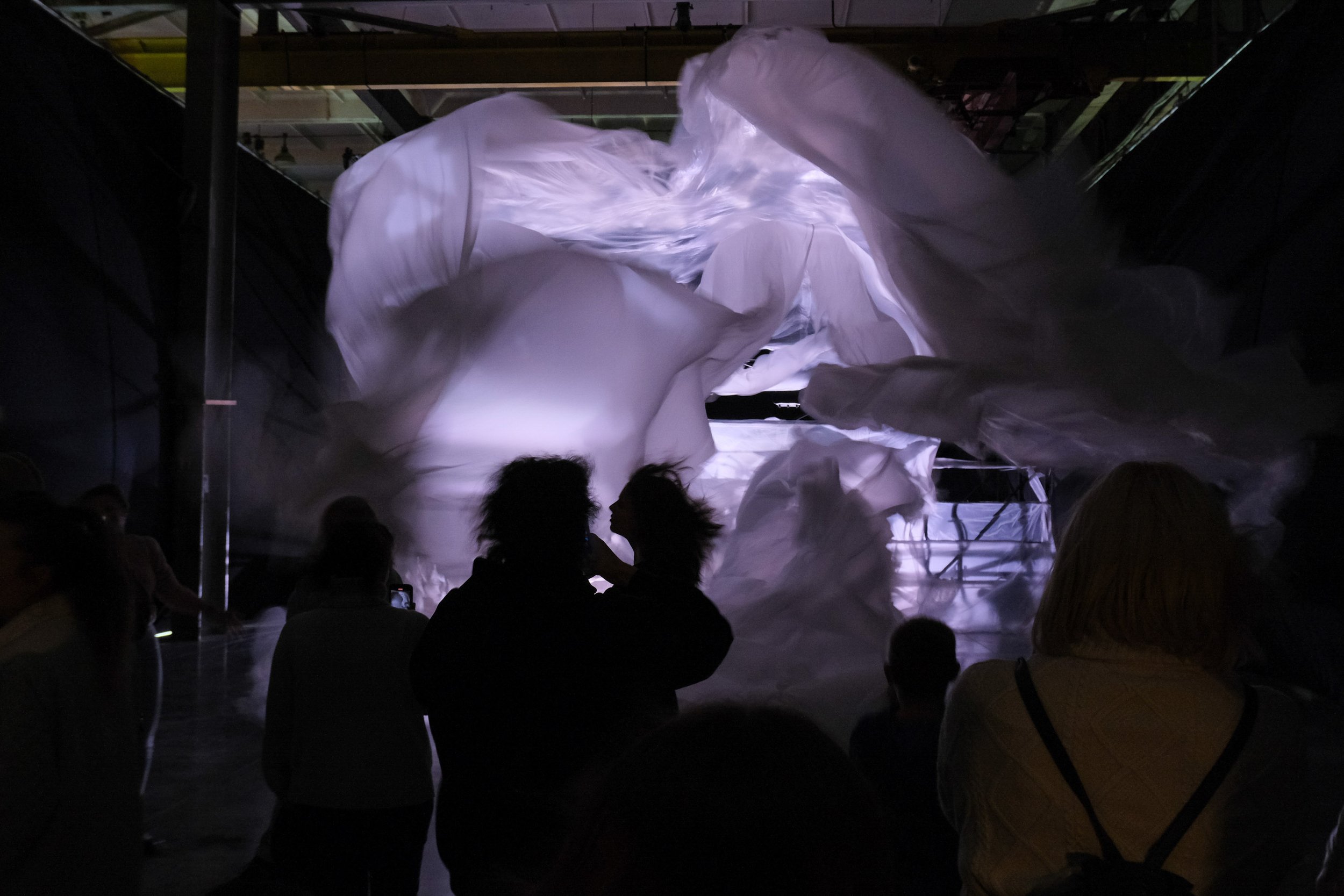 People observing large, flowing white fabric sculptures in a dark indoor space, possibly an art installation or performance involving fabric movement.