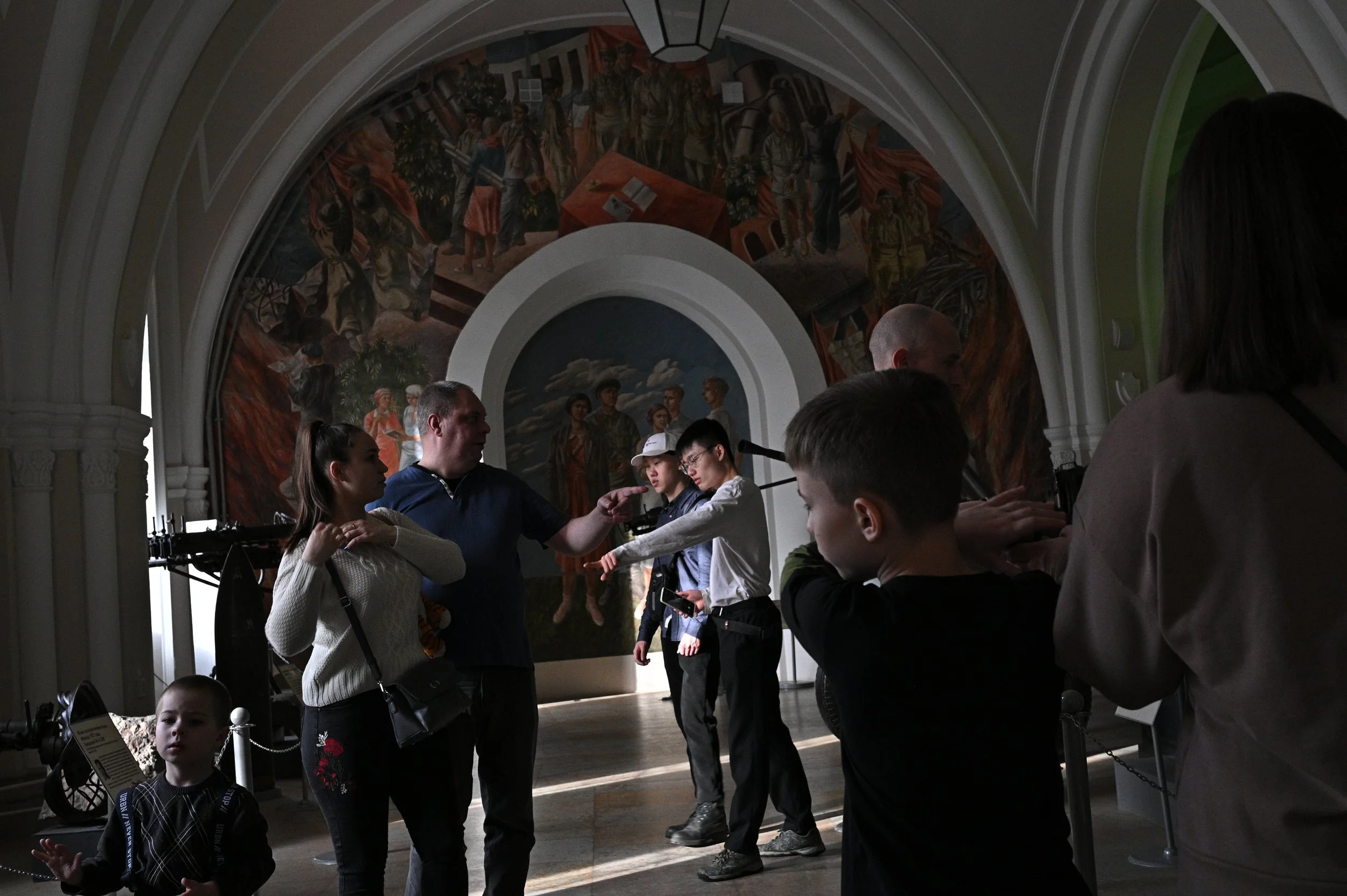 A group of people, including children and adults, inside a museum with murals on the walls. Some are observing and others are engaging in conversation.
