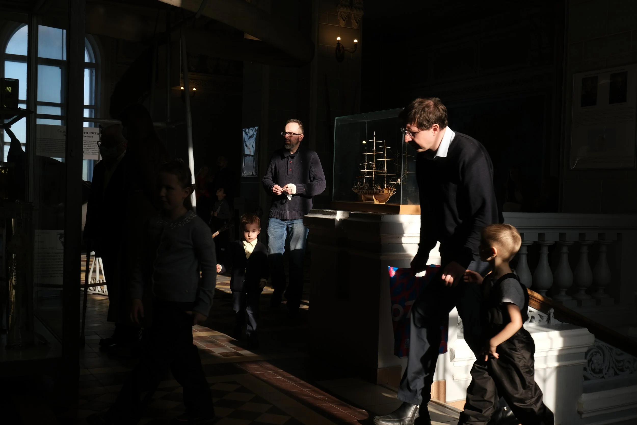 People inside a dimly lit museum exhibit area with sunlight streaming through large windows, displaying a model ship encased in glass.