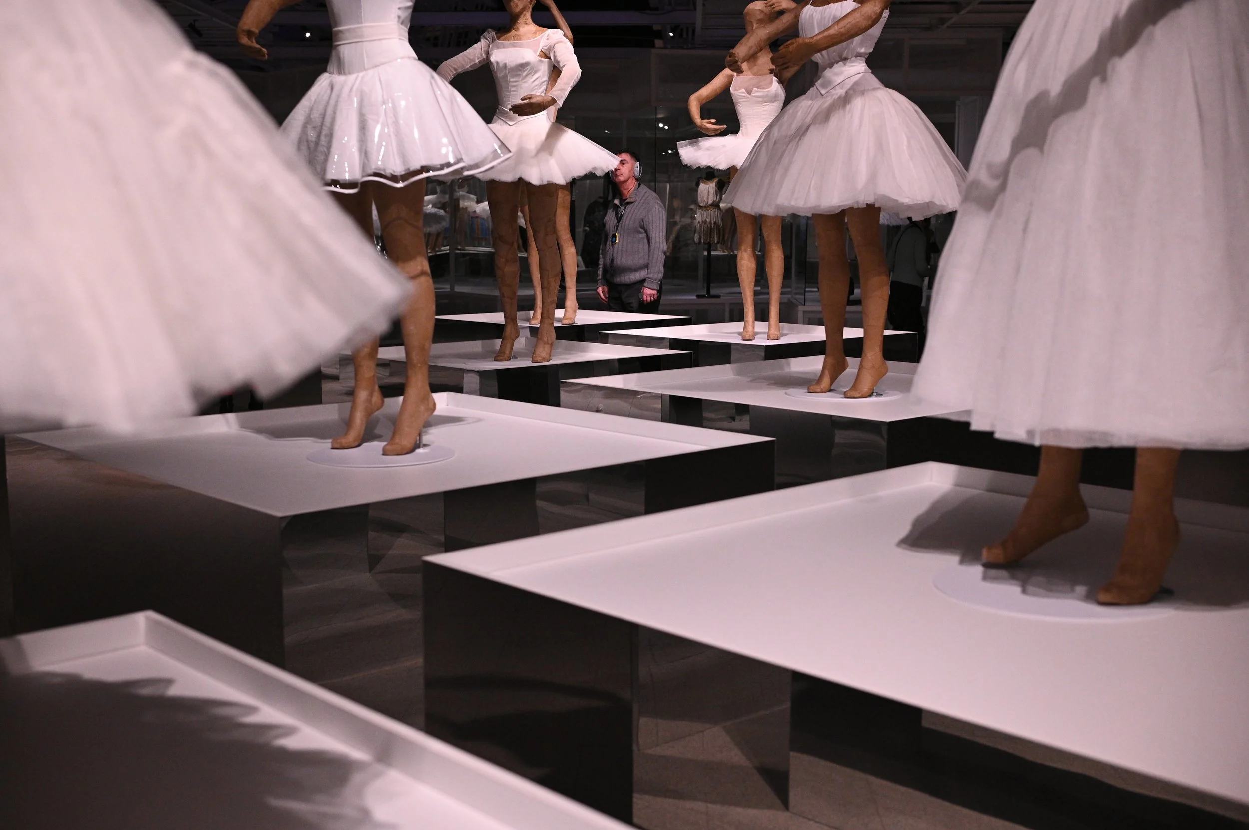 Display of mannequins dressed in white ballet tutus and pointe shoes, arranged on white platforms in a museum or gallery setting. A visitor is observing the exhibit.