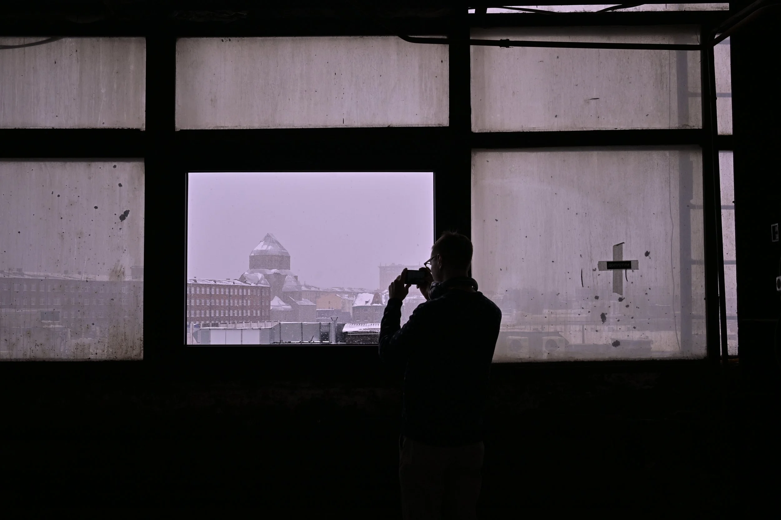 A person taking a photo of a snowy cityscape through a large window inside a dark room.