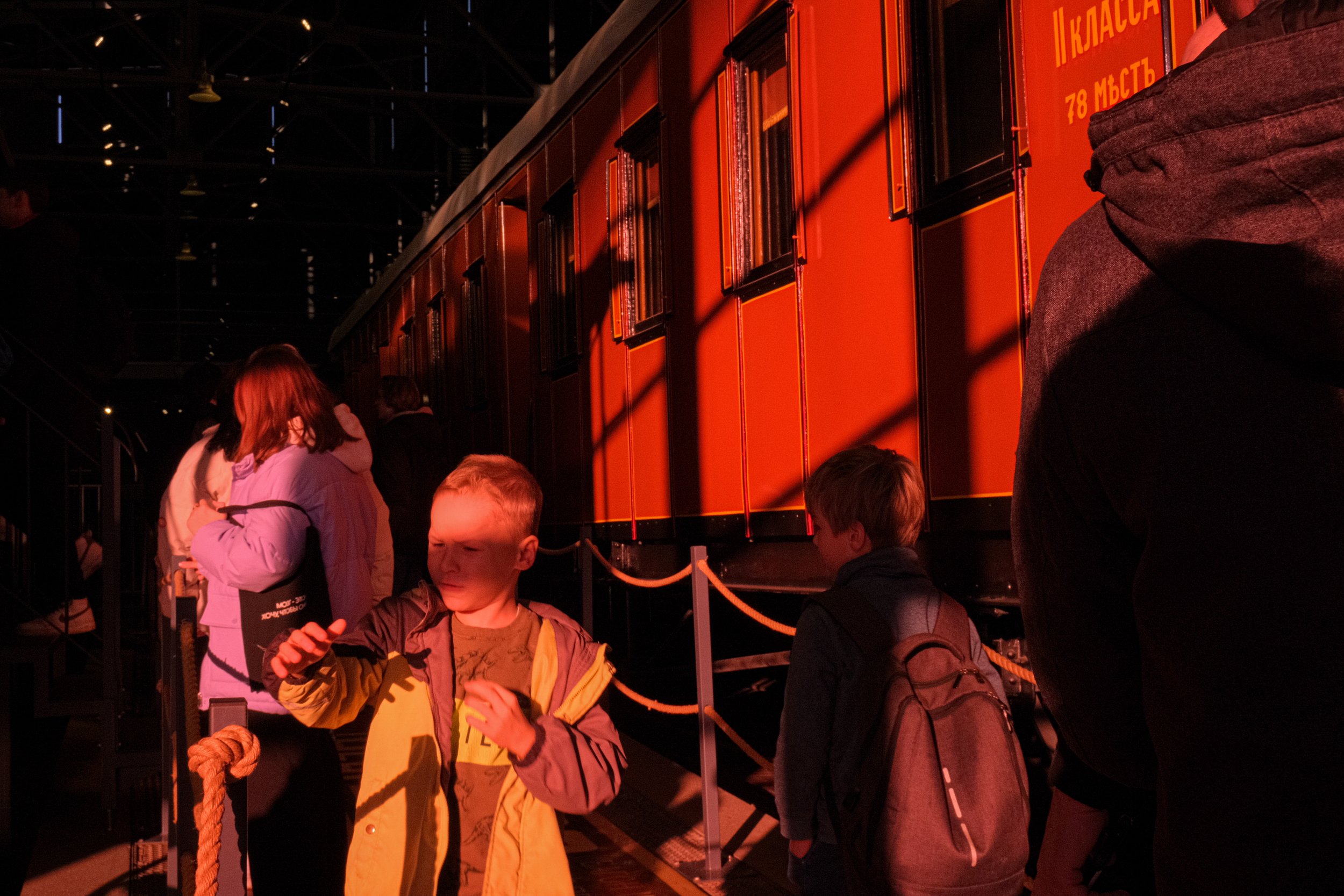 Children and adults visiting a museum exhibit with a large orange and black train display, some children are looking at their phones while others observe the train.