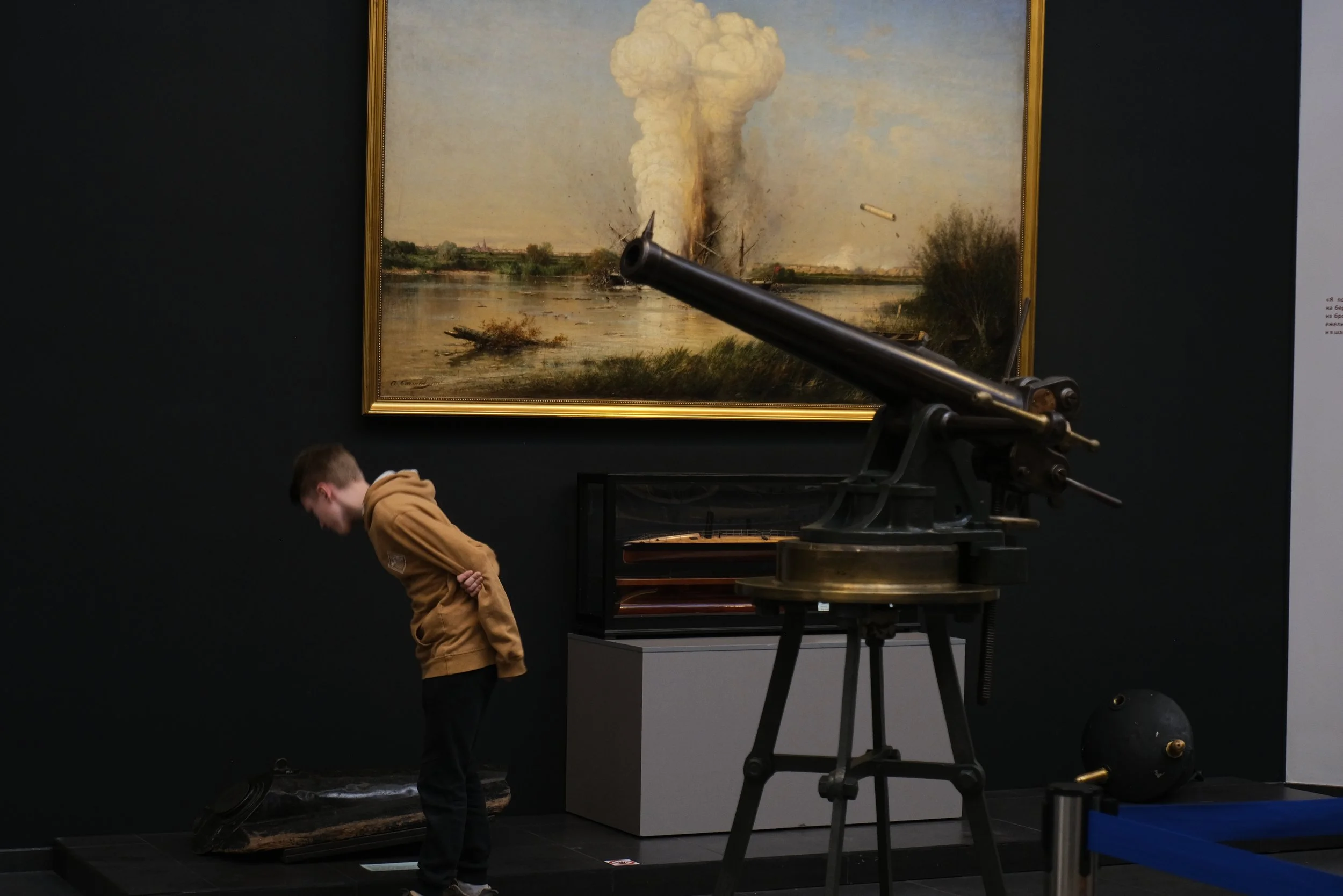 A museum exhibit featuring a large painting of a landscape with a destructive explosion, a vintage cannon, a young boy with a brown hoodie examining something on the floor, and a black sphere-shaped object on the floor to the right.