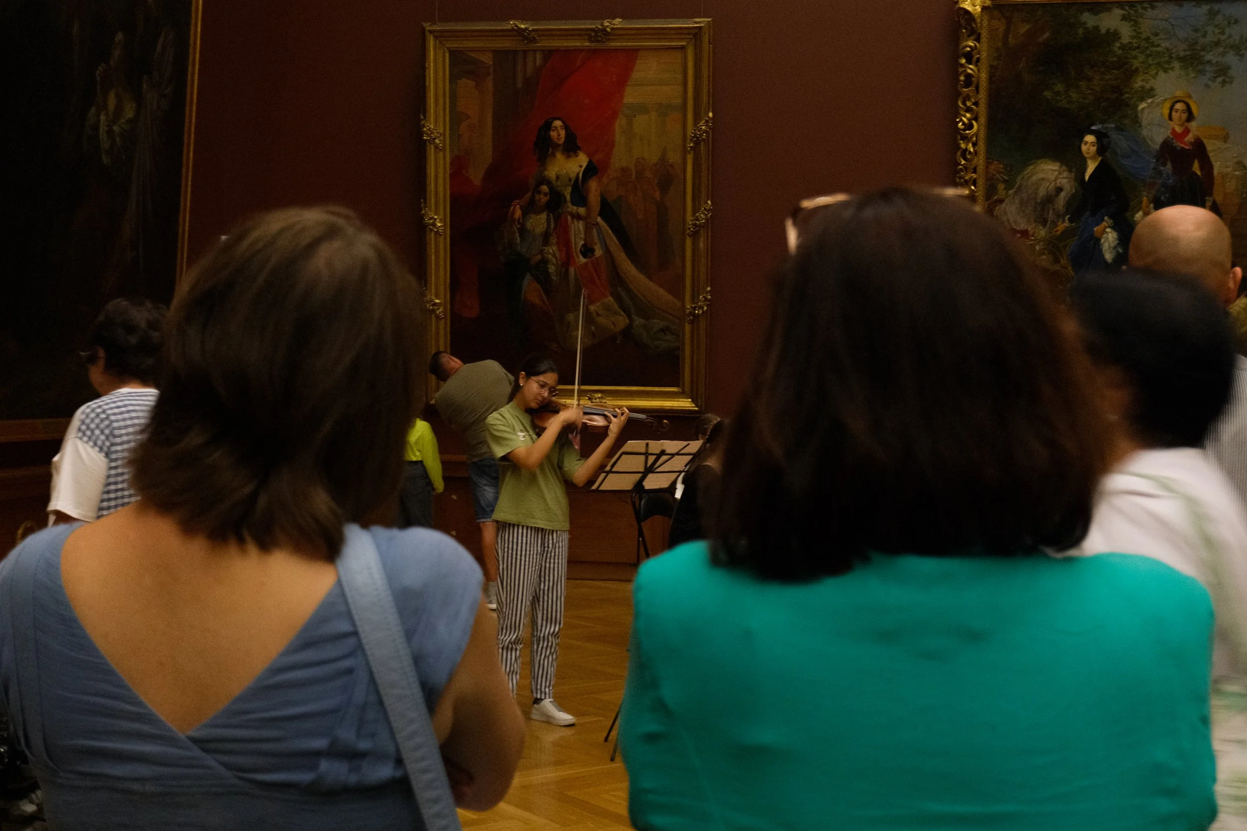 A young girl playing the violin in an art gallery, with a group of people watching her in front of large classical paintings.