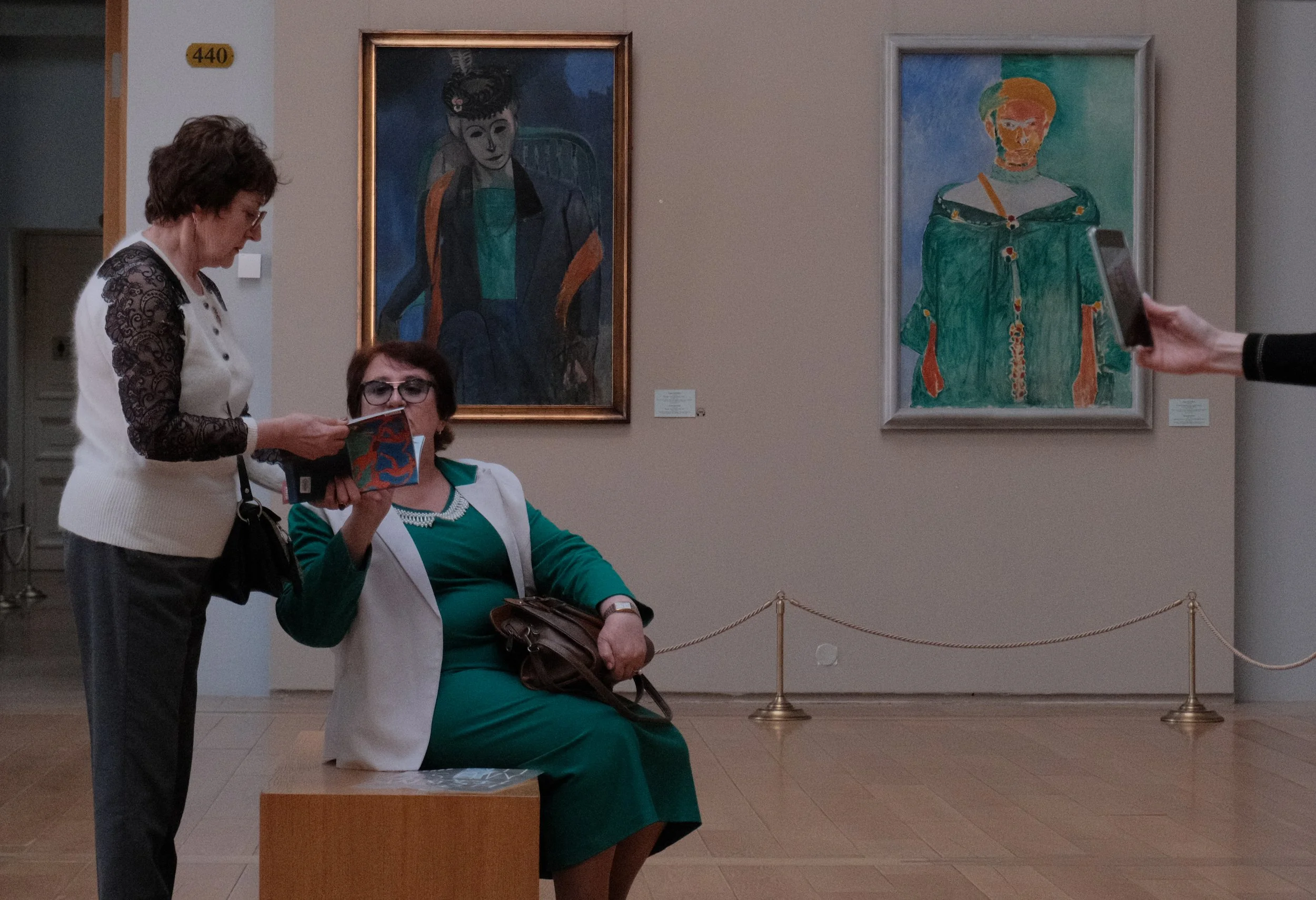 Two women are viewing artwork in a gallery. One woman, dressed in green and sitting on a bench, is holding a book or catalog. The other woman, standing next to her, is showing her something in the book. Behind them are two paintings on the wall featu