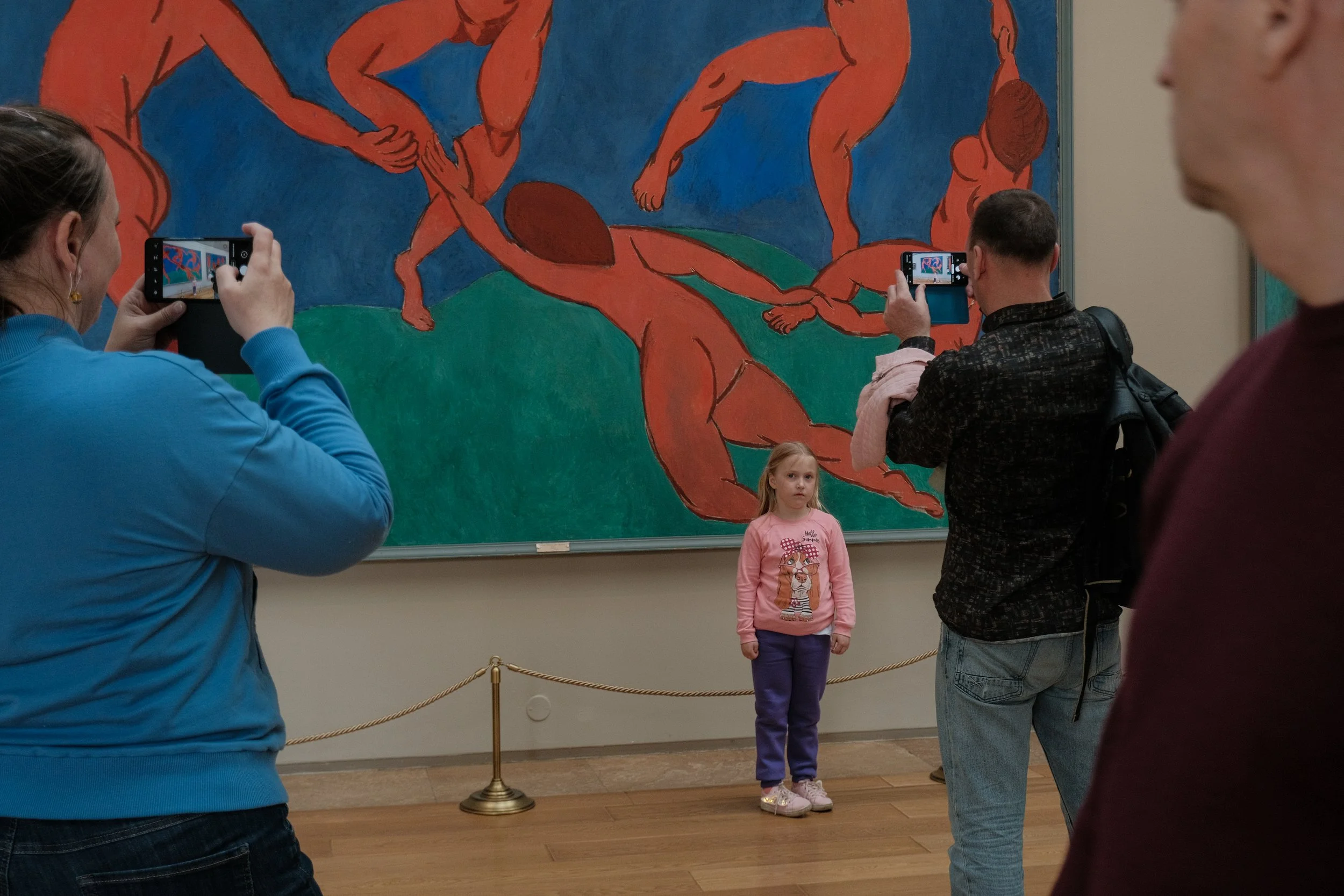 A young girl with blonde hair, wearing a pink sweater with a cat graphic, purple pants, and beige shoes, stands in front of a large painting of nude human figures in dynamic poses. Several people are taking photos of her with their phones inside an a
