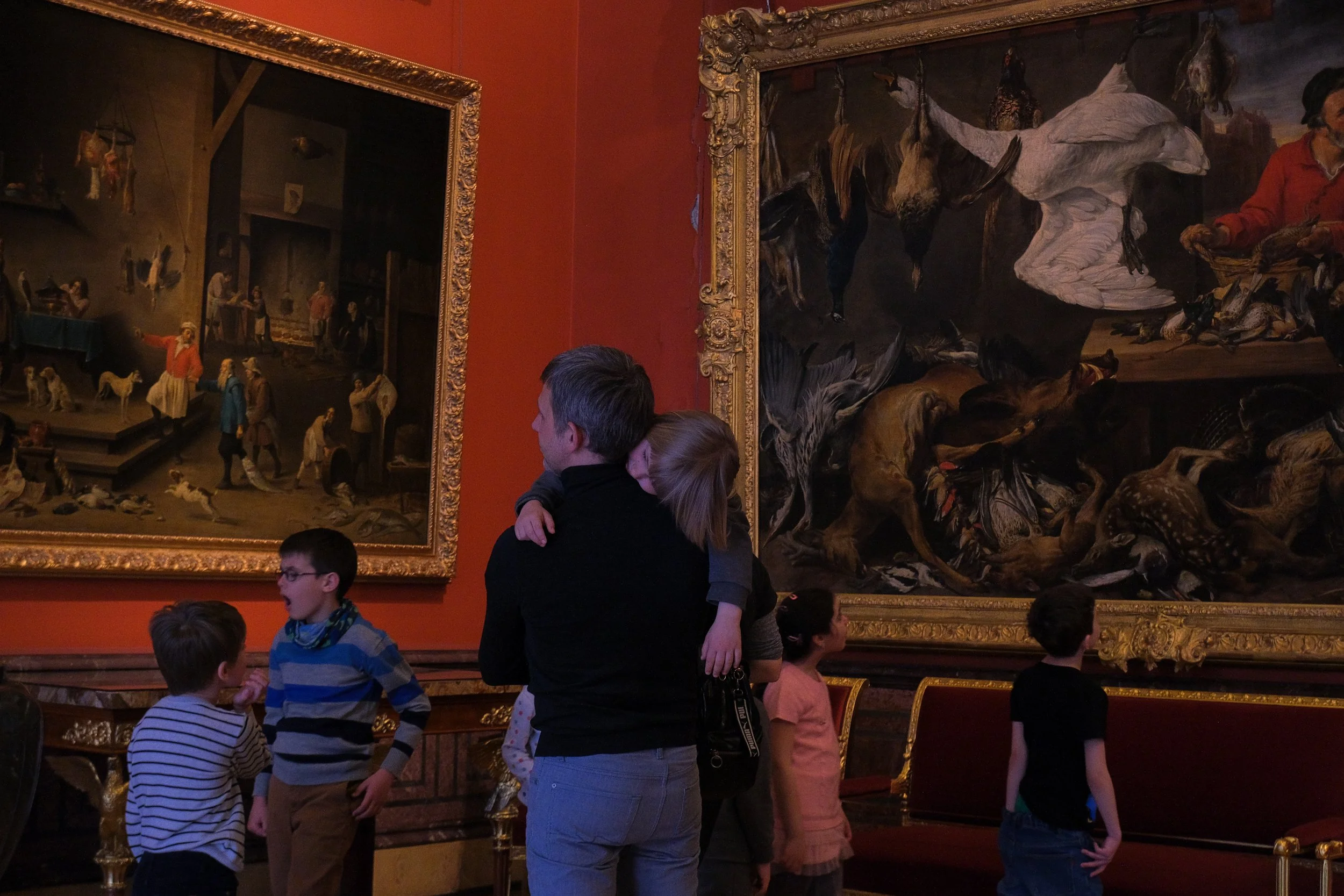 Children and adults observing large, dark paintings in an art gallery. The paintings depict historical or mythological scenes with animals and people.